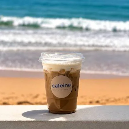 A plastic cup of iced coffee with a lid, placed on a white surface on a beach, with ocean waves in the background.