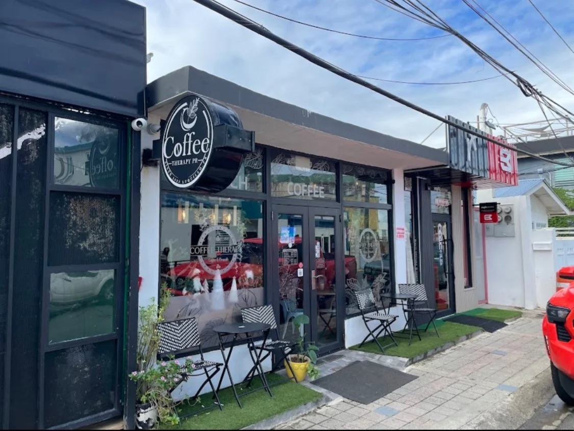 Exterior view of a coffee shop with black-framed glass windows and doors, outdoor seating with black chairs and small tables, green artificial grass mats, and a black circular sign with a coffee cup logo that reads 'Coffee Therapy'.