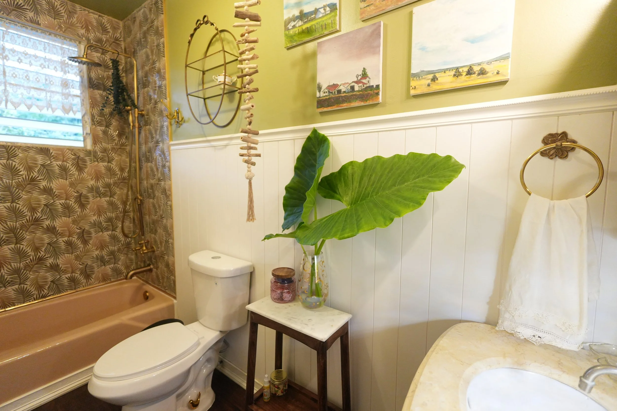A small bathroom with a shower, a toilet, and a vanity with a marble countertop. The wall above the toilet is decorated with small landscape paintings and a unique hanging seashell decoration. A large green leaf plant in a glass vase is on a small ta