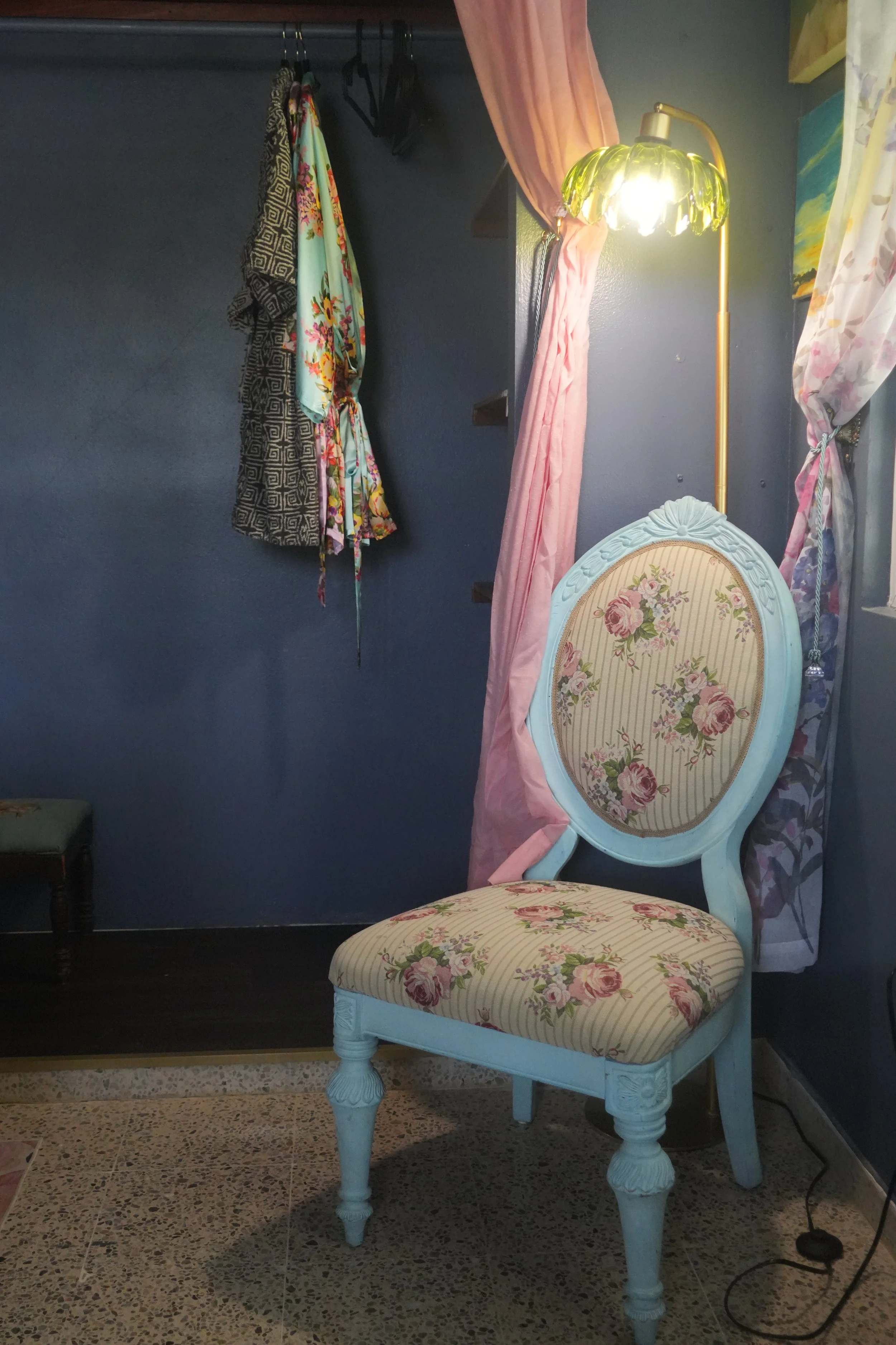 A blue and white vintage-style chair with floral upholstery in front of a dark wall. Behind the chair, there is a pink curtain tied back, with floral and patterned textiles hanging nearby. A green-colored floor lamp with a floral design is turned on,