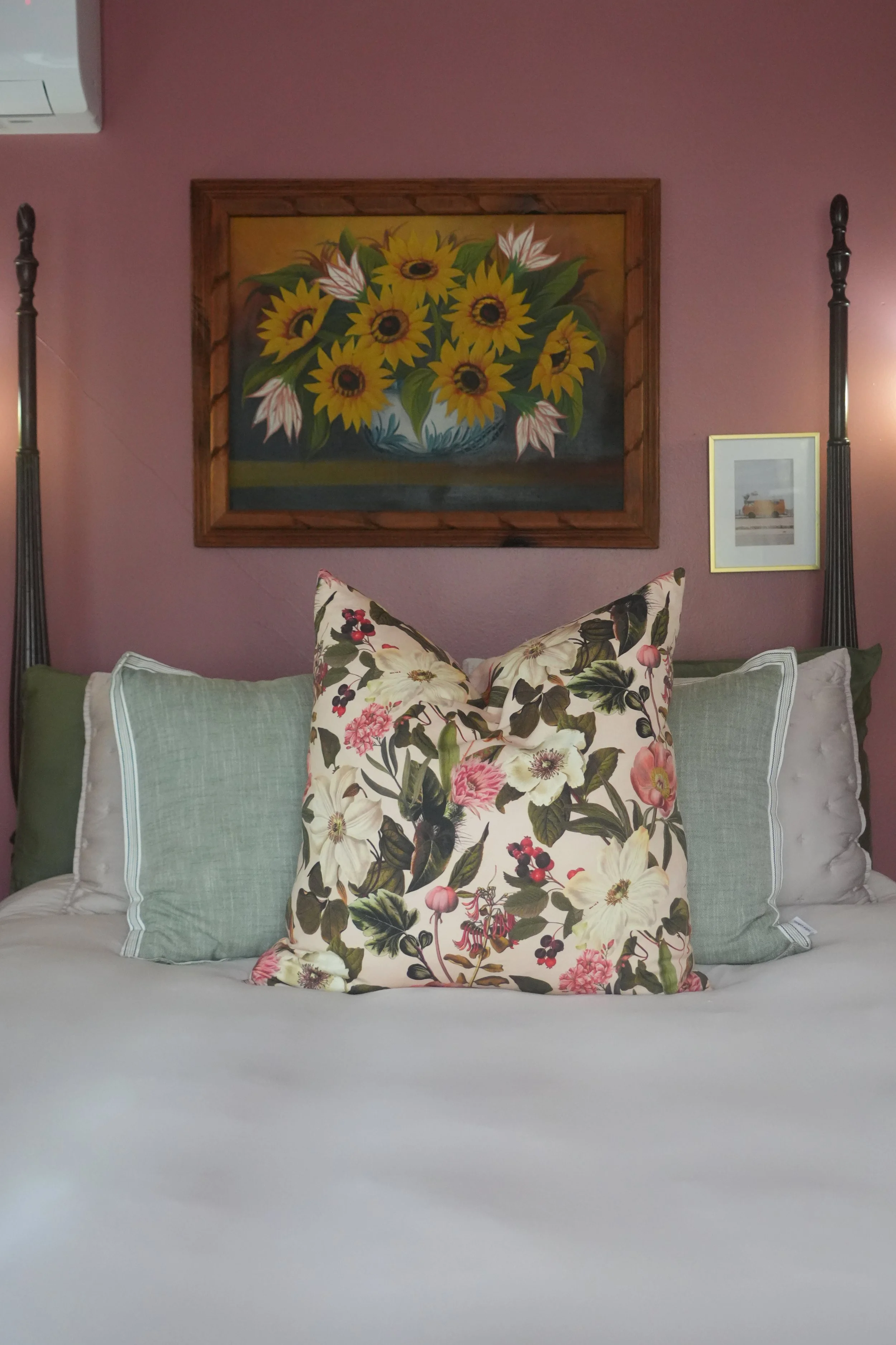 A bed with multiple pillows, including a floral patterned pillow, set against a mauve wall with framed artwork of sunflowers and a picture of a vintage van.