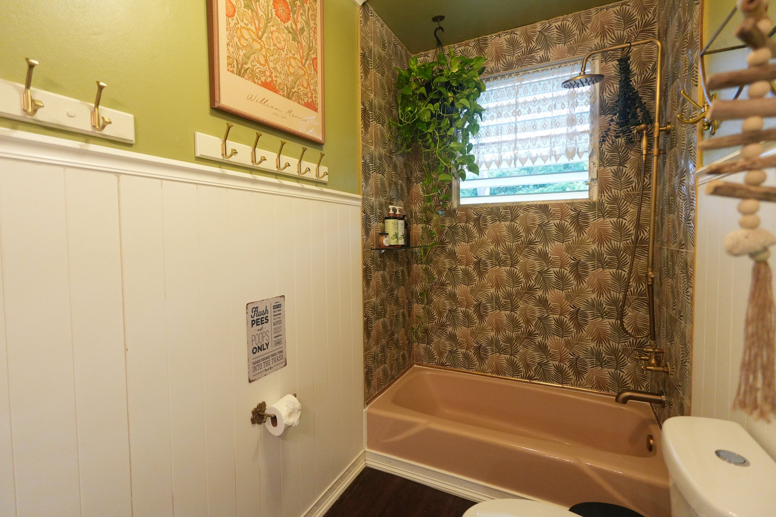A bathroom with a glass block window, tropical leaf-patterned shower tile, a tall green plant, and gold-colored fixtures, including a shower head and towel hooks, and a framed William Morris artwork on a green wall.