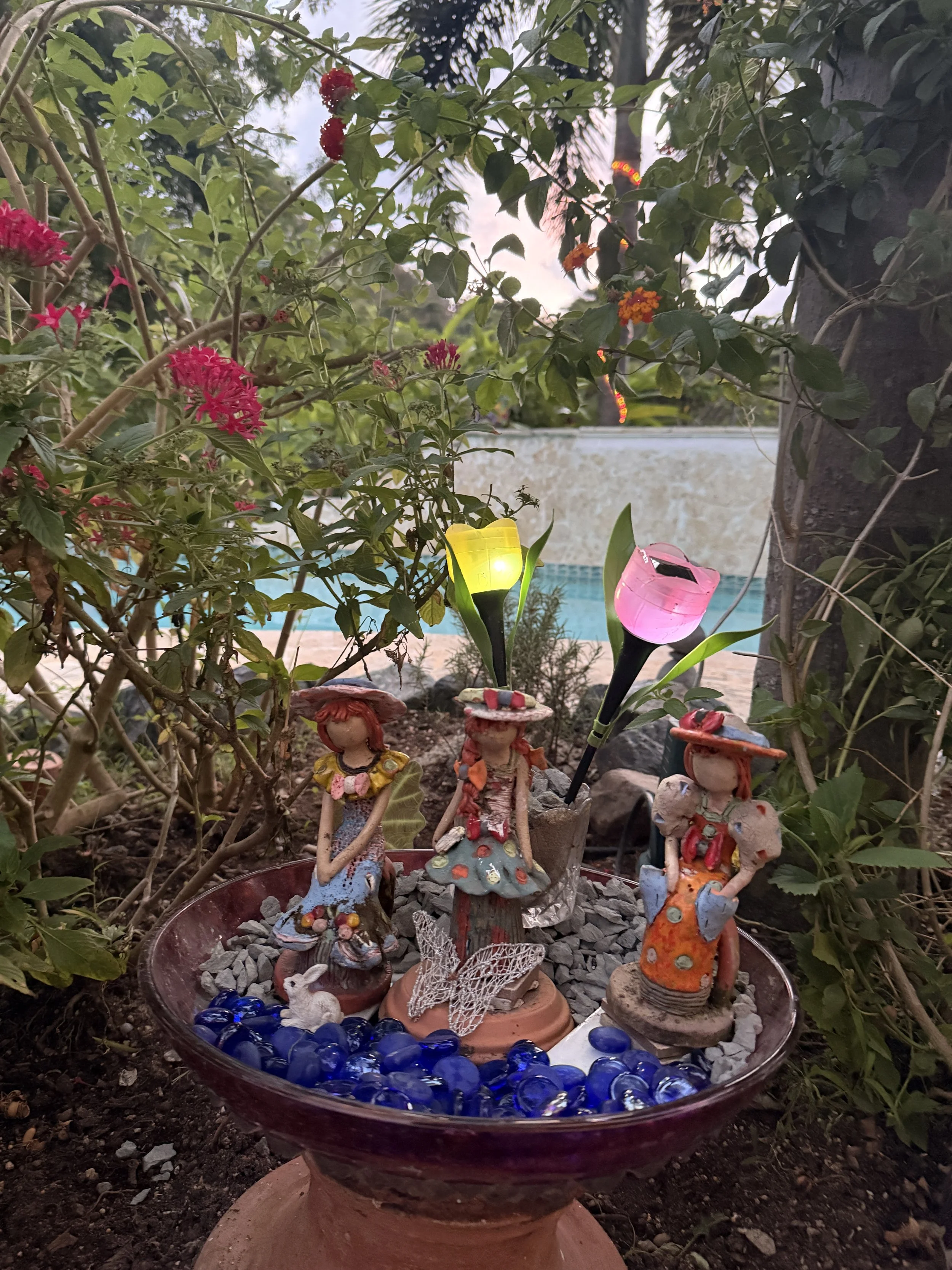Small decorative figurines of children and fairies placed in a garden bowl filled with blue glass stones, surrounded by greenery and flowers with two colorful lanterns nearby.