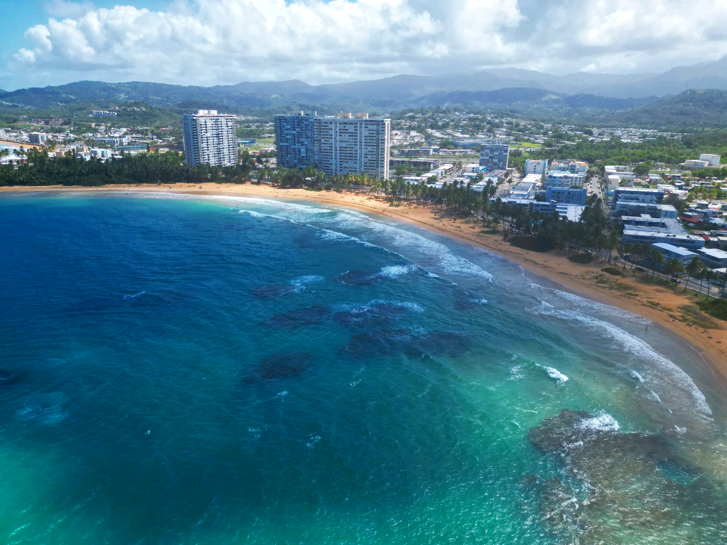 Aerial view of la Costa Azul with clear blue water, sandy shore, palm trees, high-rise buildings, and a city surrounded by green hills and mountains in the background.