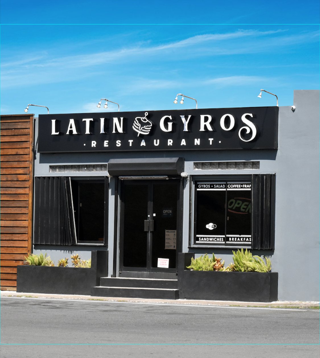 storefront of Latin Gyros restaurant with black sign and white lettering, menu signs on windows, and outdoor plants, under a blue sky.