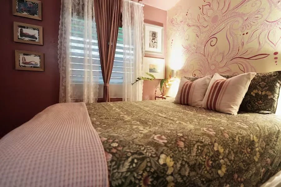 A cozy bedroom with a bed decorated with floral bedspread and multiple pillows, pink walls with wall art, curtains covering windows, and a soft light source illuminating the room.