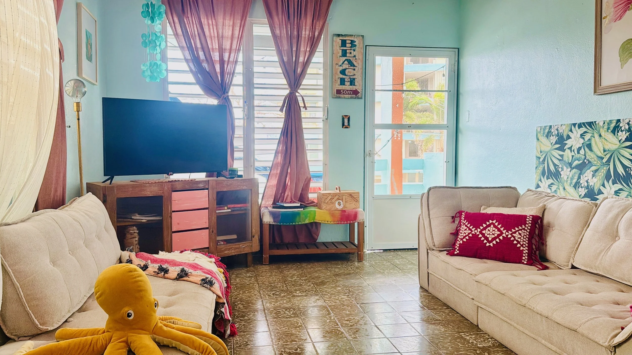 Living room with two beige sofas, a colorful blanket, a yellow octopus plush toy, and a red pillow. There is a flat-screen TV on a wooden stand, a colorful table, pink curtains, and a door and window revealing a balcony with tropical plants outside.
