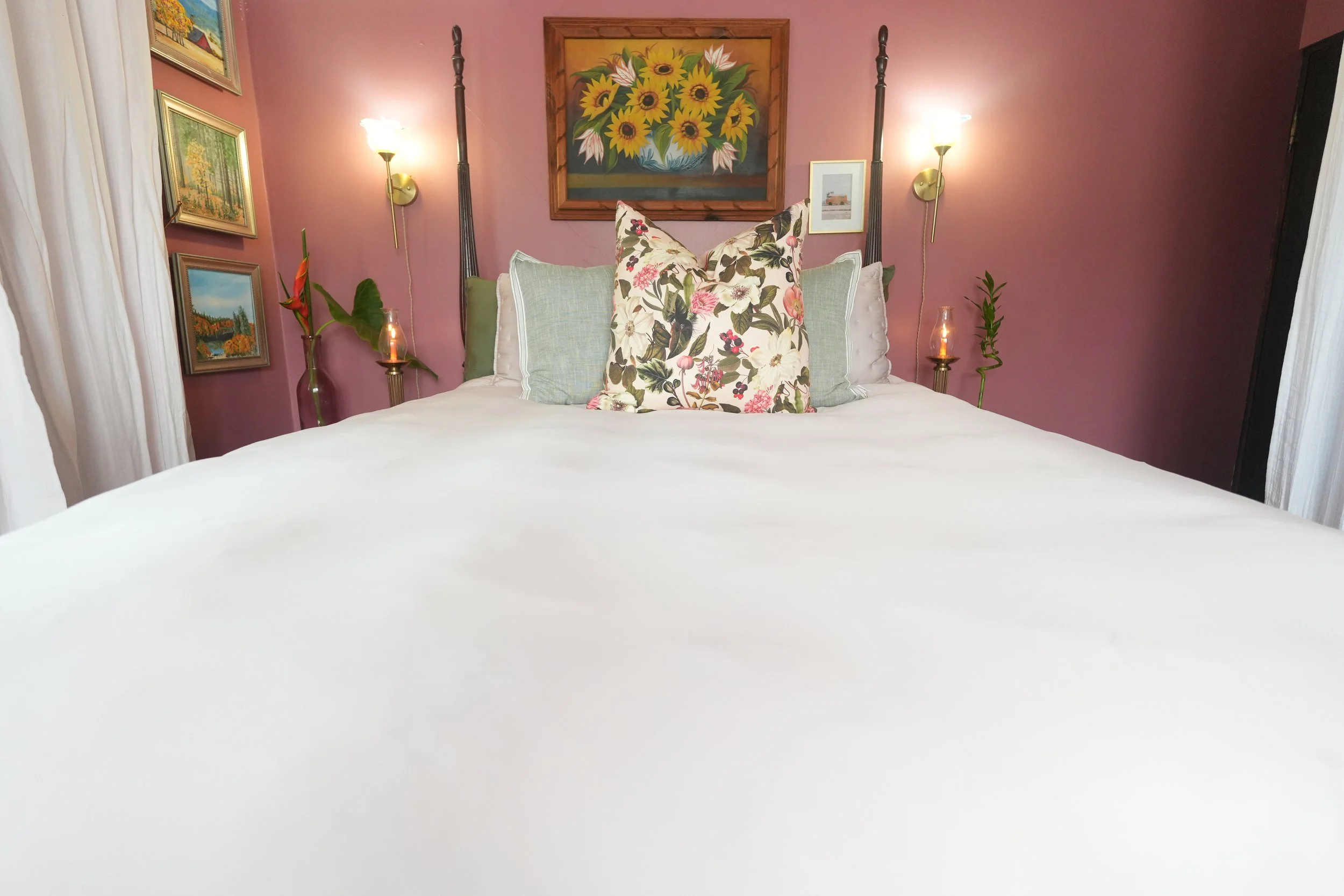 A bedroom with a pink wall, decorated with framed landscape paintings and a large painting of sunflowers. There is a bed with white bedding, accented by floral and light green pillows. On either side of the bed, there are small tables with vases hold