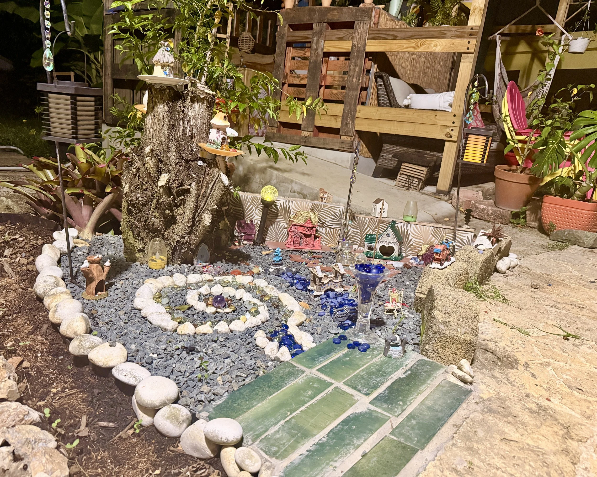 Nighttime outdoor scene featuring a small shrine with miniature houses, surrounded by garden decorations, rocks, and plants. A wooden structure in the background has a bed with white bedding. Various hanging ornaments and garden accessories are visible.