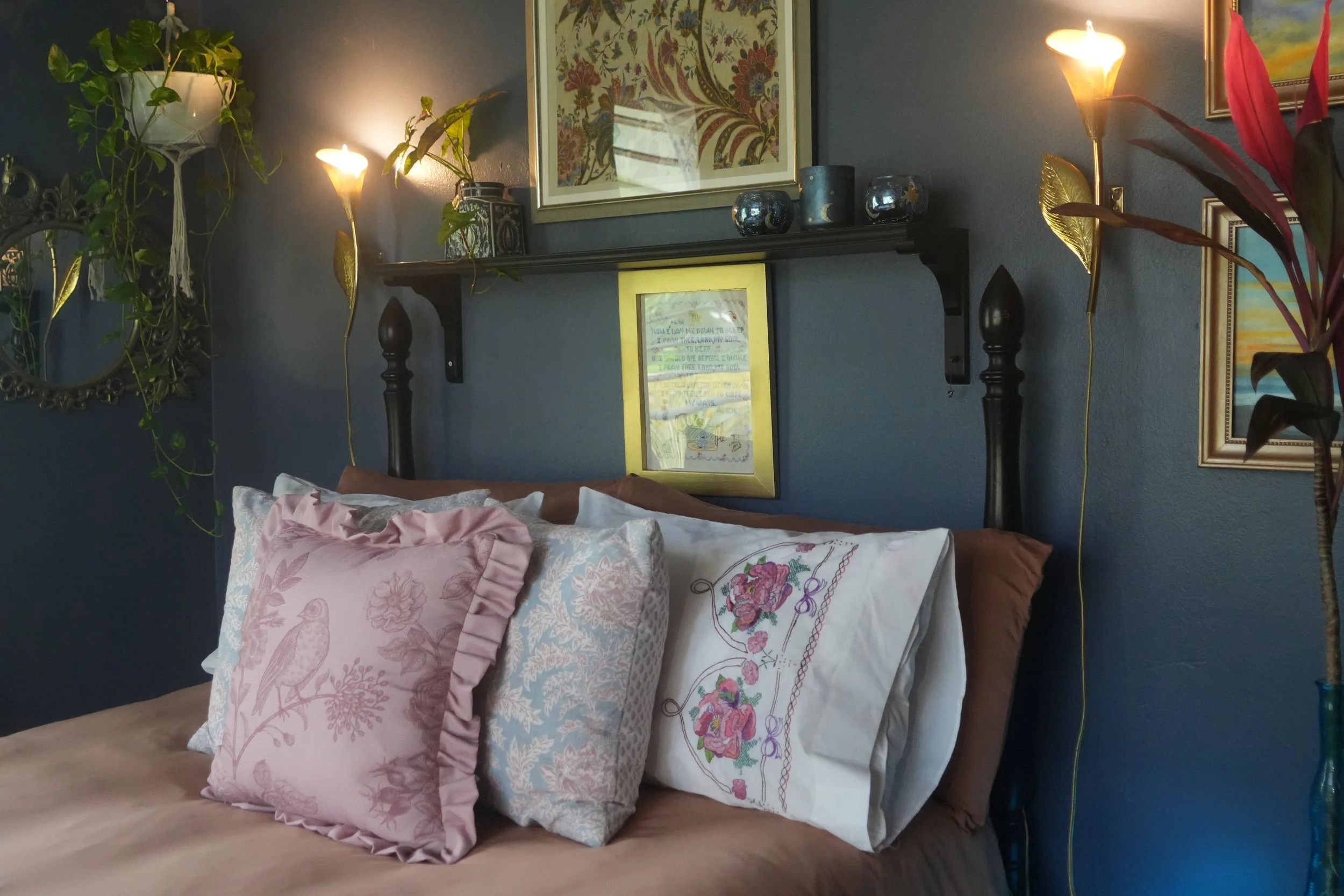 A bedroom with a dark blue wall, decorated with framed artwork and a black shelf holding decorative items. There are two table lamps and a tall plant on each side of the bed, which has floral and embroidered pillows and bedding. A framed drawing or p