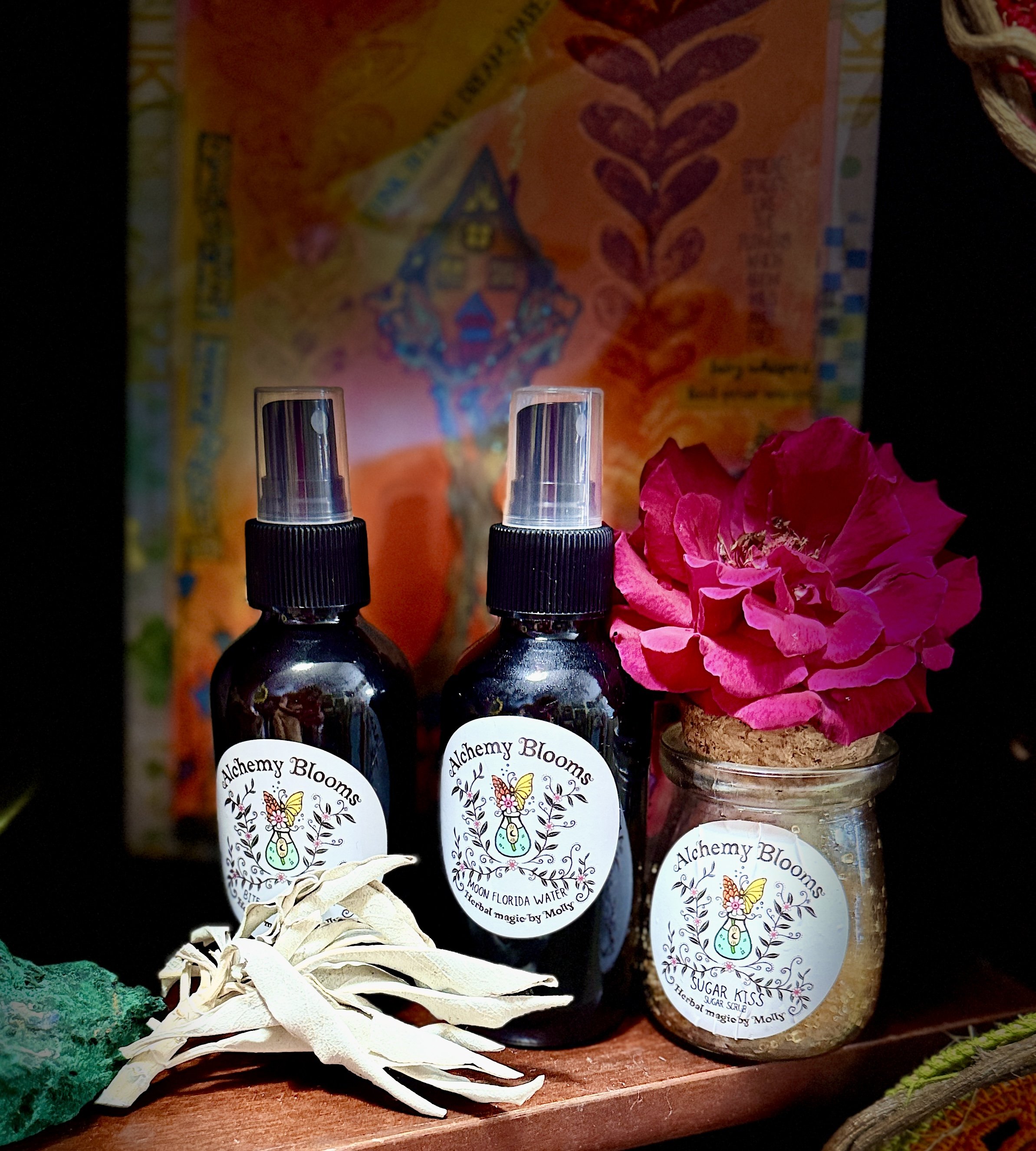 Two small bottles of Alchemy Blooms herbals with plant-themed labels, one labeled 'Sugar Kiss,' and a small jar with a similar label, set on a wooden surface with decorative items including a dried flower and a green sponge.