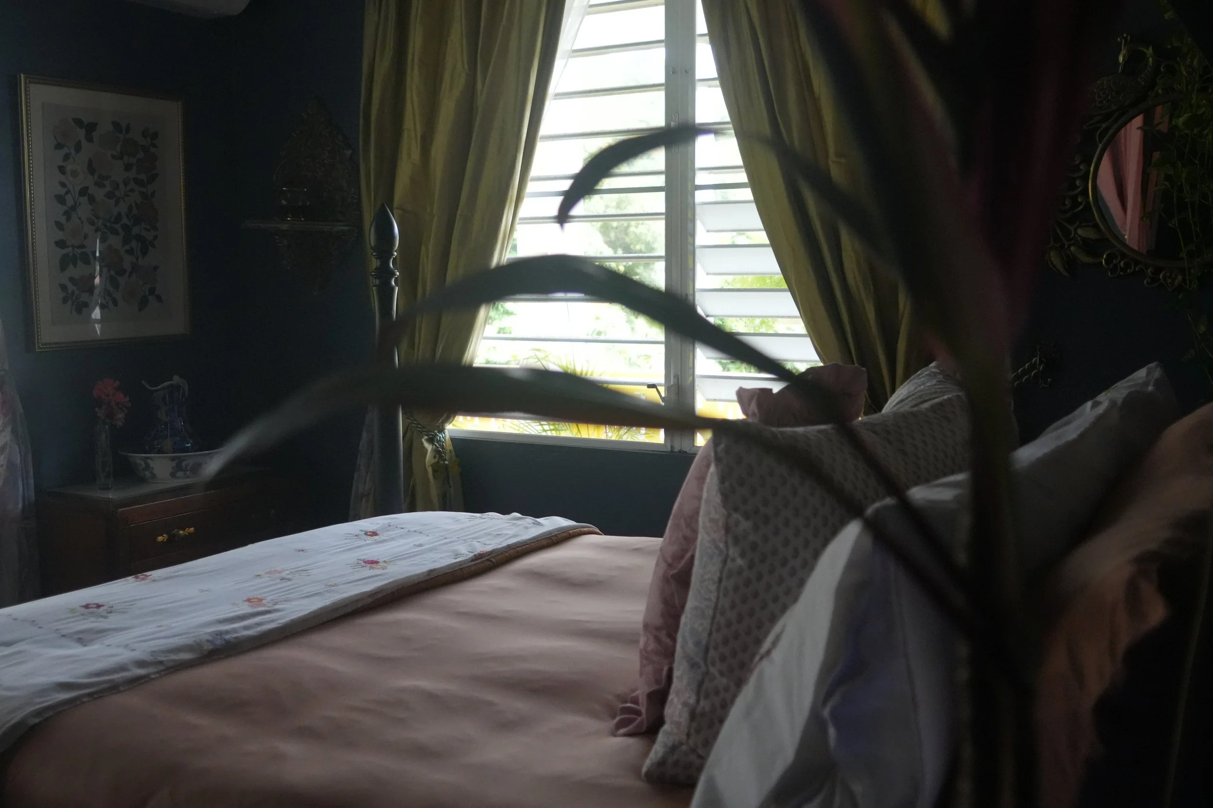 A bedroom with a large window with yellow curtains, a bed with a floral bedspread, a framed picture of leaves on the wall, and a bedside table with a glass vase and a decorative item inside.