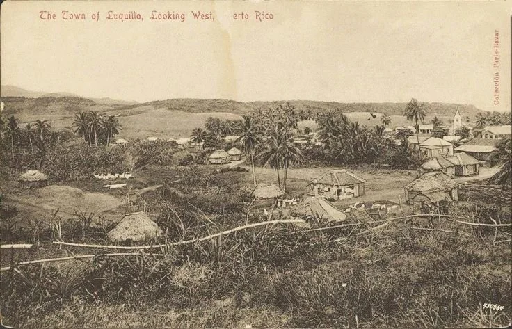 A historic black and white photograph of the town of Luquillo, Puerto Rico, with small houses, palm trees, and hilly landscape in the background.