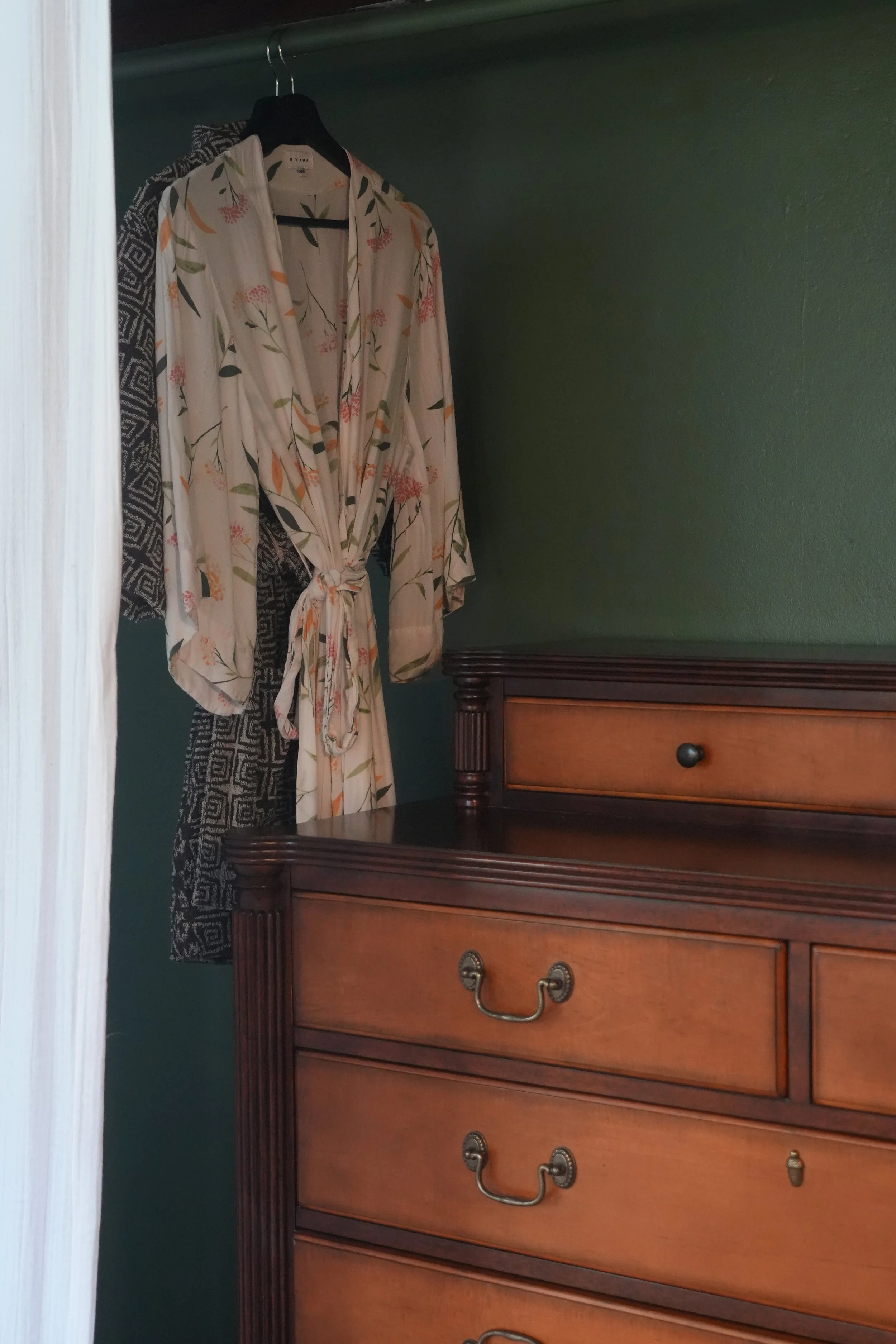 A wooden dresser with three drawers and brass handles, placed against a dark green wall. A floral robes hangs on a black hanger inside a closet with a dark green interior, partially visible from an open door.