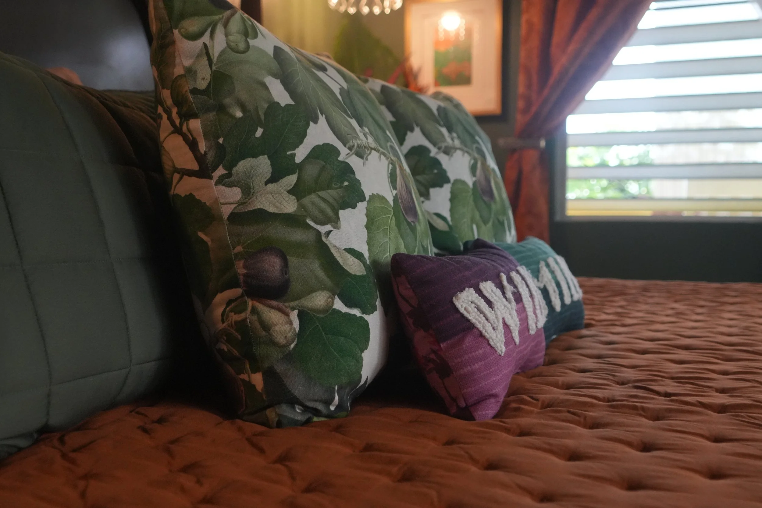 Close-up of decorative pillows on a brown textured couch, featuring a leafy green patterned pillow and a purple pillow with the word 'LOVE' embroidered in white. In the background, outdoor light filters through white blinds, and interior wall art is 