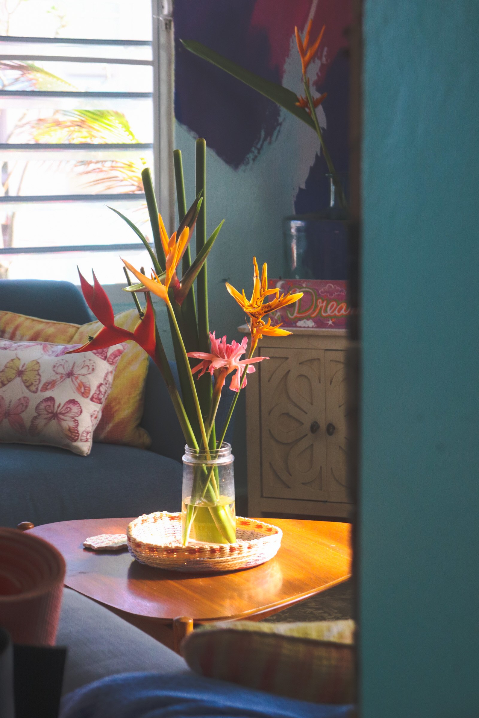 A bouquet of orange and pink flowers in a glass vase on a wooden table with a woven placemat. A window with blinds can be seen in the background, with a blue wall and a colorful picture behind the plants.