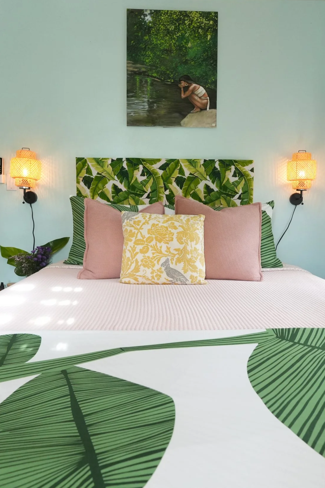 A bedroom with pastel green walls, a headboard with a tropical leaf pattern, and various decorative pillows featuring botanical and bird prints. There are two wall-mounted lamps with woven shades on either side of the bed, and a framed picture of a person crouched by a stream hanging above the headboard.