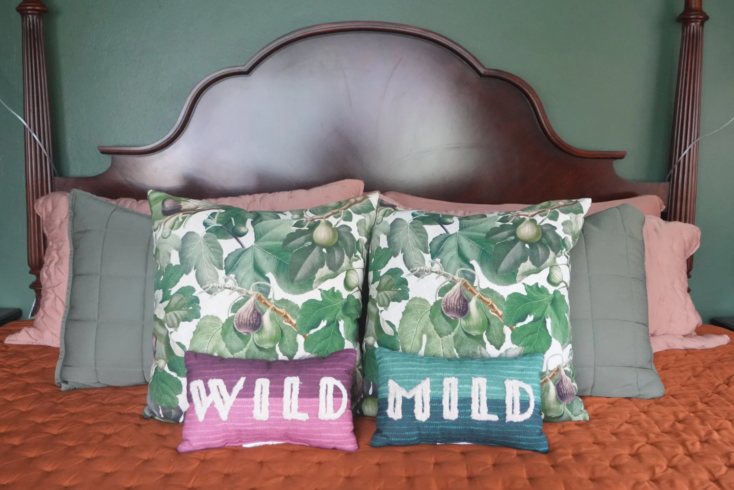 A bed with a green wooden headboard and orange quilted bedspread, decorated with pillows, including one with a fig tree pattern and two smaller pillows with the words 'wild child' and 'wild mind'.
