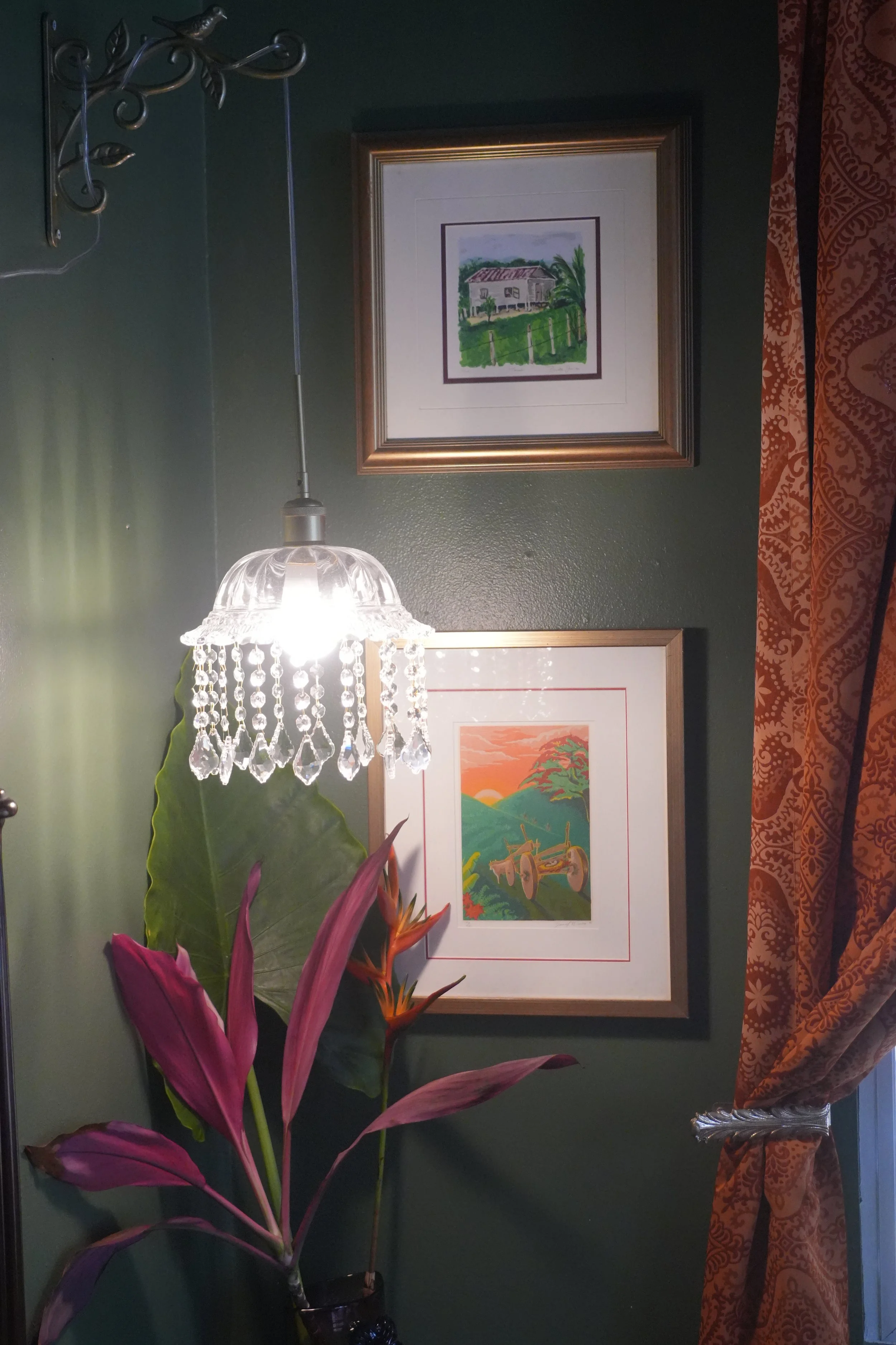 Interior wall decorated with framed artwork, a crystal chandelier, and a large pink and green plant in the foreground. The artwork depicts a rural scene with a house and greenery.