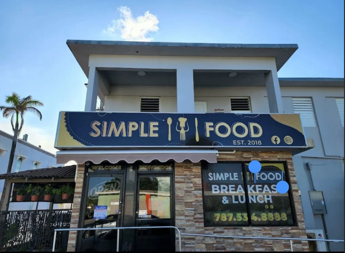Front view of a restaurant named Simple Food, established in 2018, serving breakfast and lunch. Sign indicates its focus on simple meals with cutlery icons. The building has a stone facade and a glass door with a welcome sign. There are small potted plants on a shelf to the left of the entrance, and a palm tree is visible on the left side of the image.