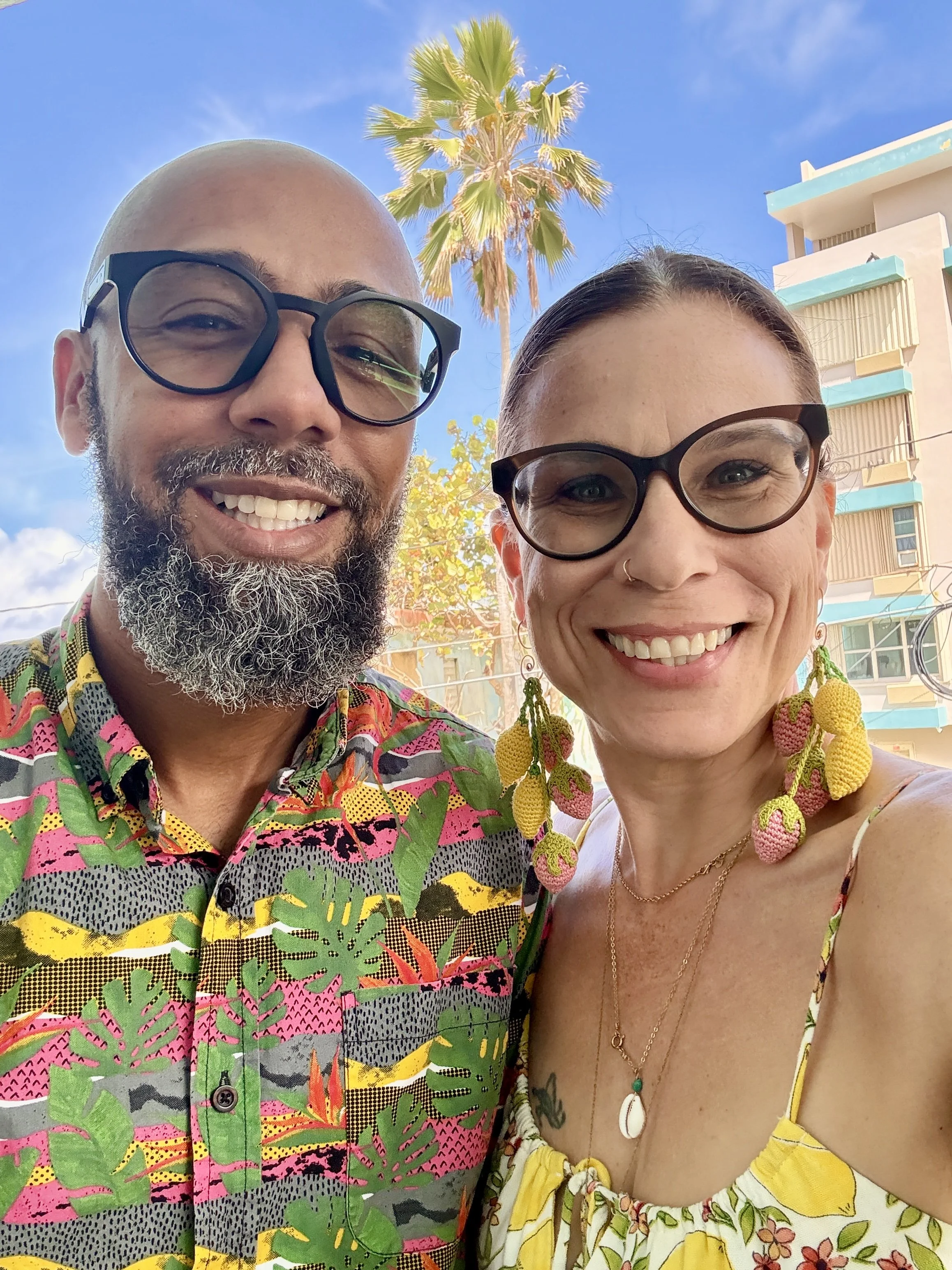A smiling man with glasses and a beard standing next to a smiling woman with glasses, both wearing colorful, tropical-patterned clothing, outdoors with a palm tree and a building in the background.
