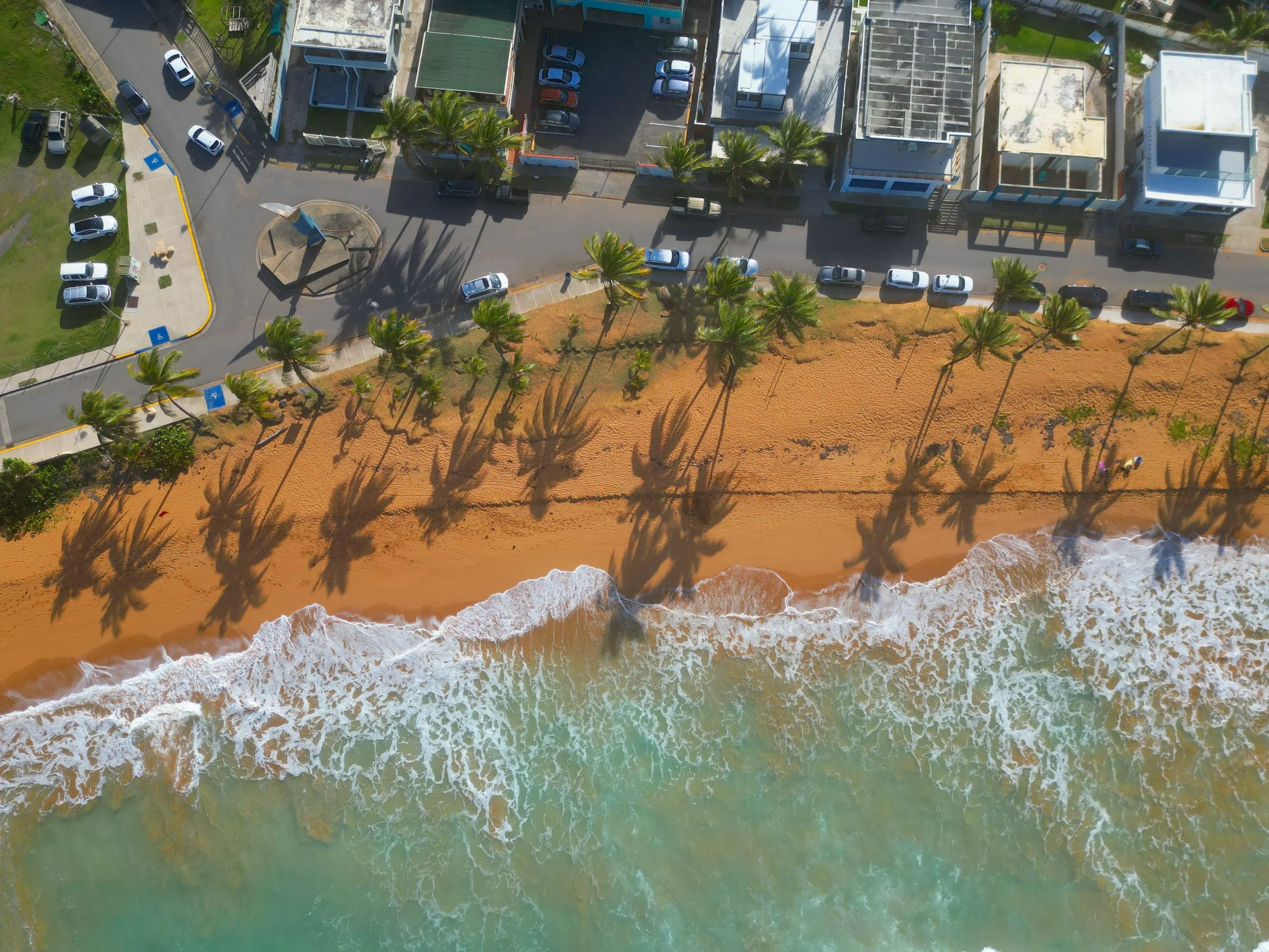 Aerial view of the street beside La Playa Pared beach with palm trees, parked cars, and modern buildings.