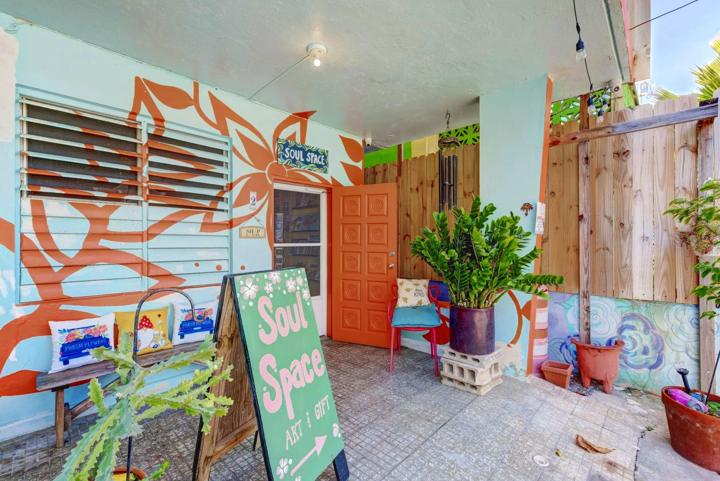 The exterior of SoulSpace, a colorful outdoor art space with a large orange floral mural on the wall, a door painted orange, and various plants in pots. There are boutique signs for art, and gifts, along with decorative pillows and chairs.