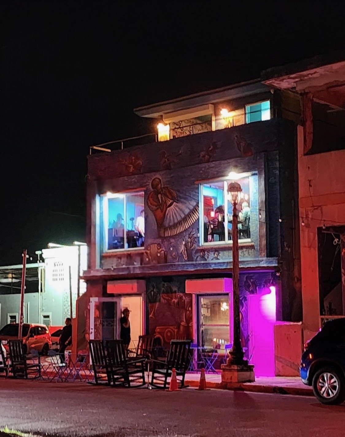 A colorful building with murals, neon lights, and people inside at night, with chairs and a streetlamp outside.