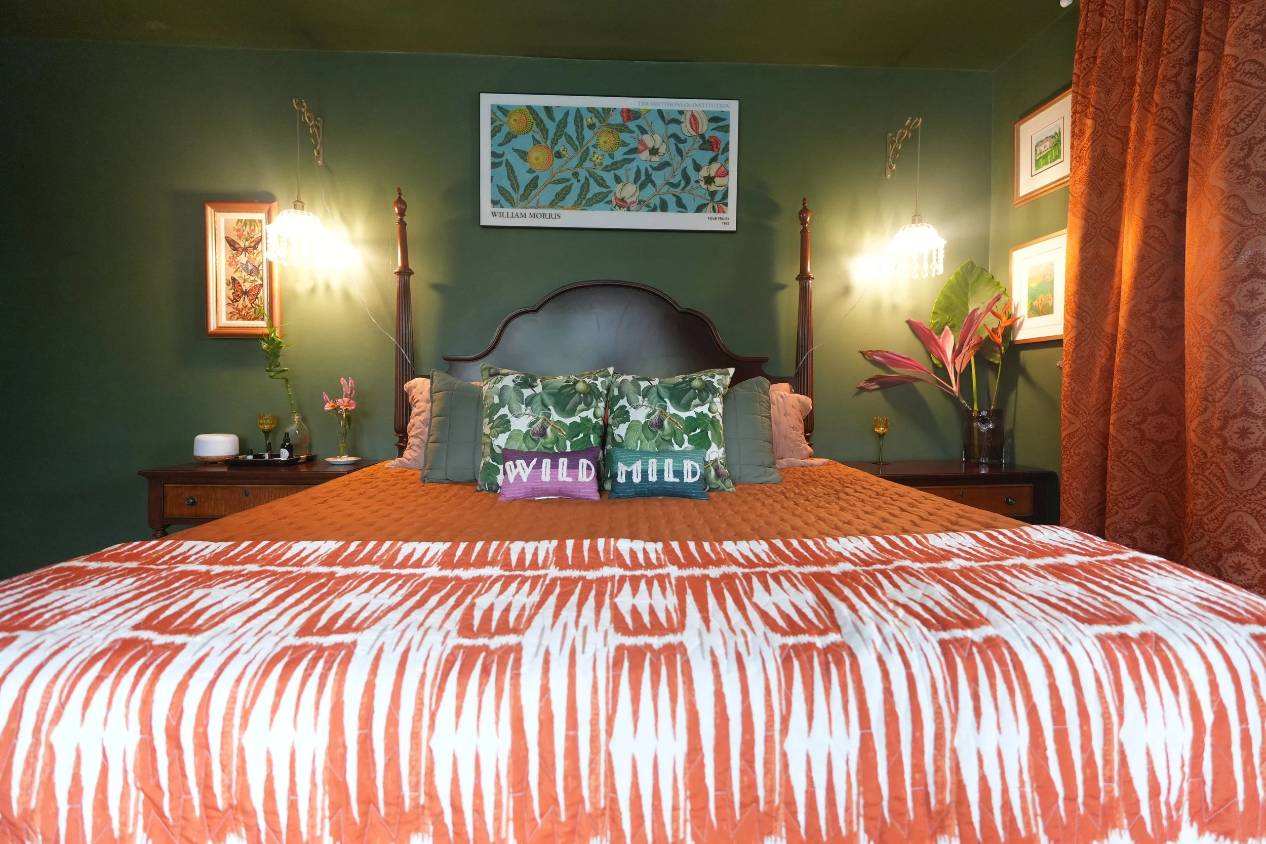A cozy bedroom with green walls and framed artwork, decorated with tropical plants, featuring a bed with patterned throw pillows that read "WILD" and "MILD," and a wooden nightstand with a white diffuser and a glass of wine.