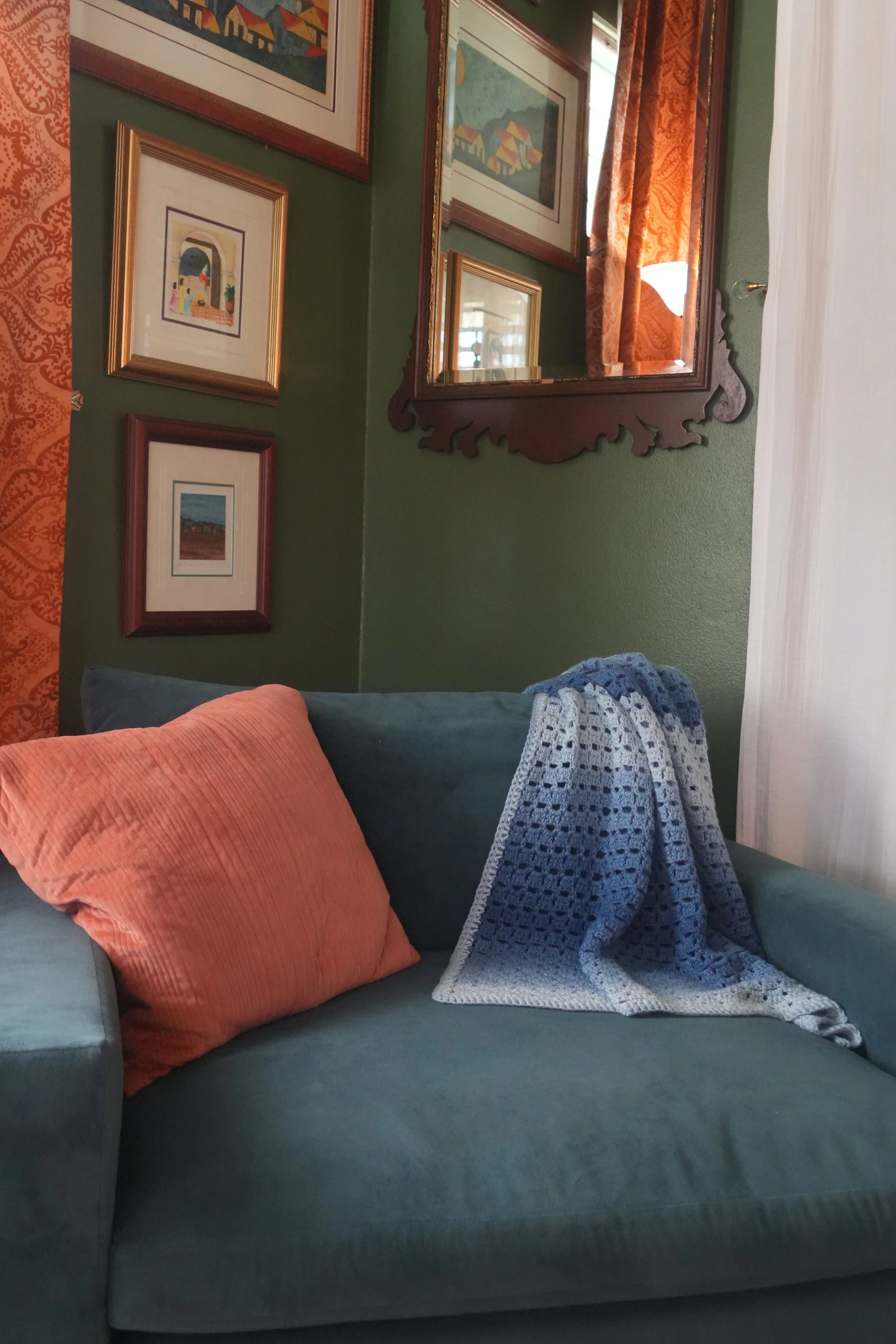 A living room with framed artwork on green walls, a mirror with a wooden carved frame, a sofa with orange and dark cushions, and a blue crochet blanket draped over the sofa.