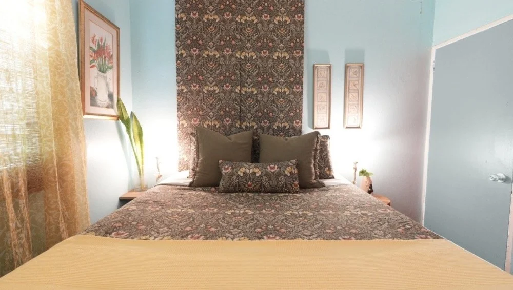 A cozy bedroom with a bed featuring a patterned bedspread and pillows, floral artwork on the left wall, and a decorative tapestry behind the headboard. There are two bedside tables with small lamps, and a window with sheer curtains on the left side.