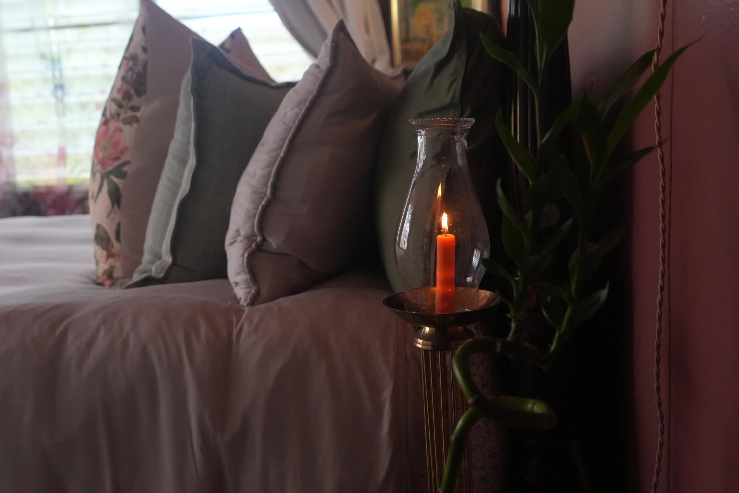 A cozy indoor scene featuring a bed with decorative pillows, a glass oil lamp with a lit candle, and a tall green leafy plant next to a pink wall, with a window in the background.