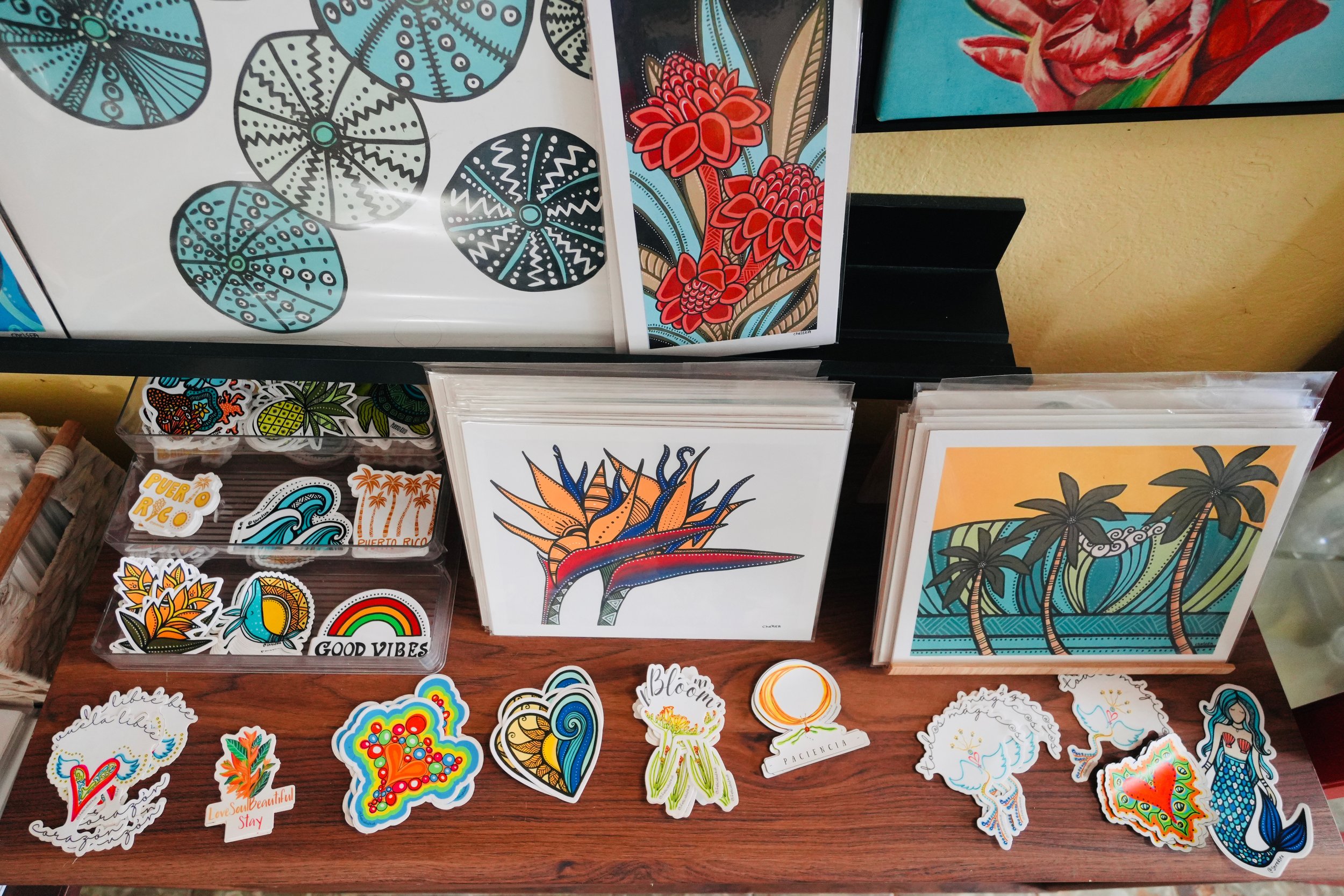 Colorful artwork and stickers with tropical and nature themes, including flowers, leaves, ocean waves, and rainbows, displayed on a table and wall.