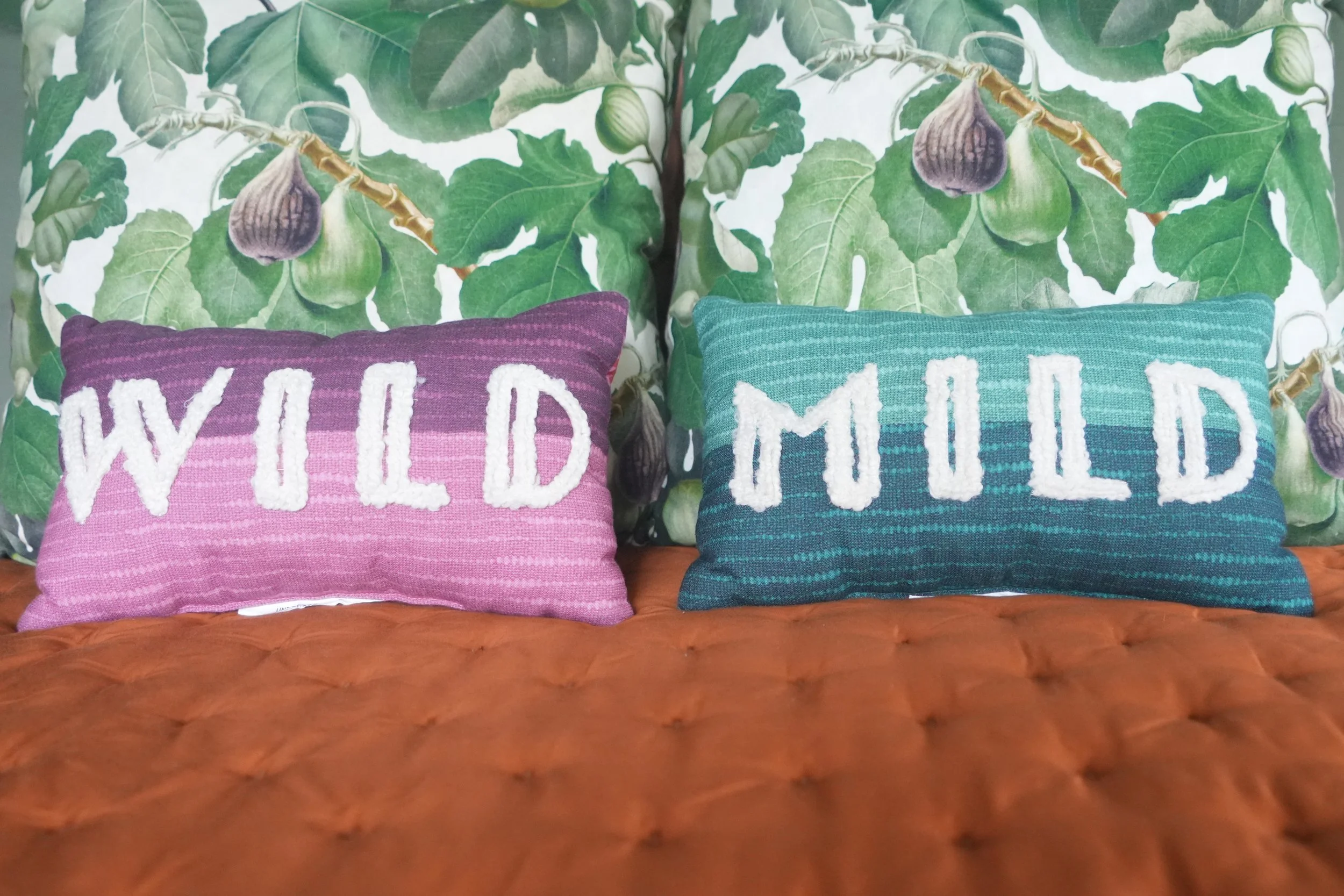 Two decorative pillows resting on a tufted leather couch; one pink with white embroidered text that says 'WILD', and one teal with white embroidered text that says 'MIND.' Behind them, there is a wallpaper with a green foliage pattern and figs.