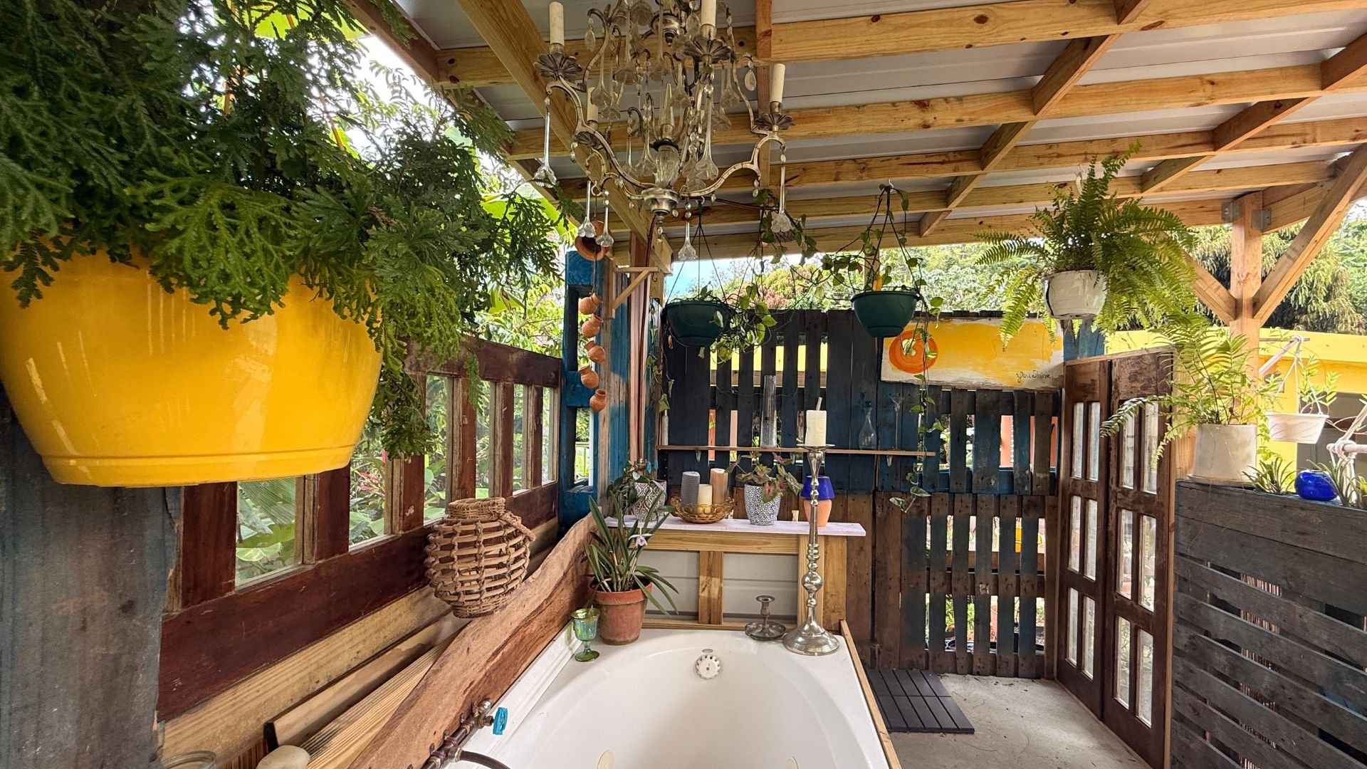 A cozy outdoor spa or hot tub area with a wooden fence, hanging plants, and decorative items like candles and a chandelier, surrounded by lush greenery.