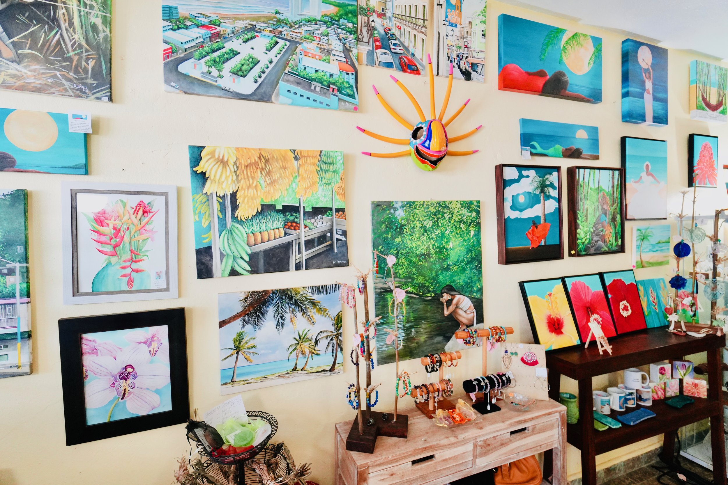 Art gallery wall with various colorful paintings depicting tropical scenes, city streets, and nature. A handmade mask with bright colors and tentacle-like appendages is mounted on the wall. Display tables below hold jewelry, crafts, and small decorative items.