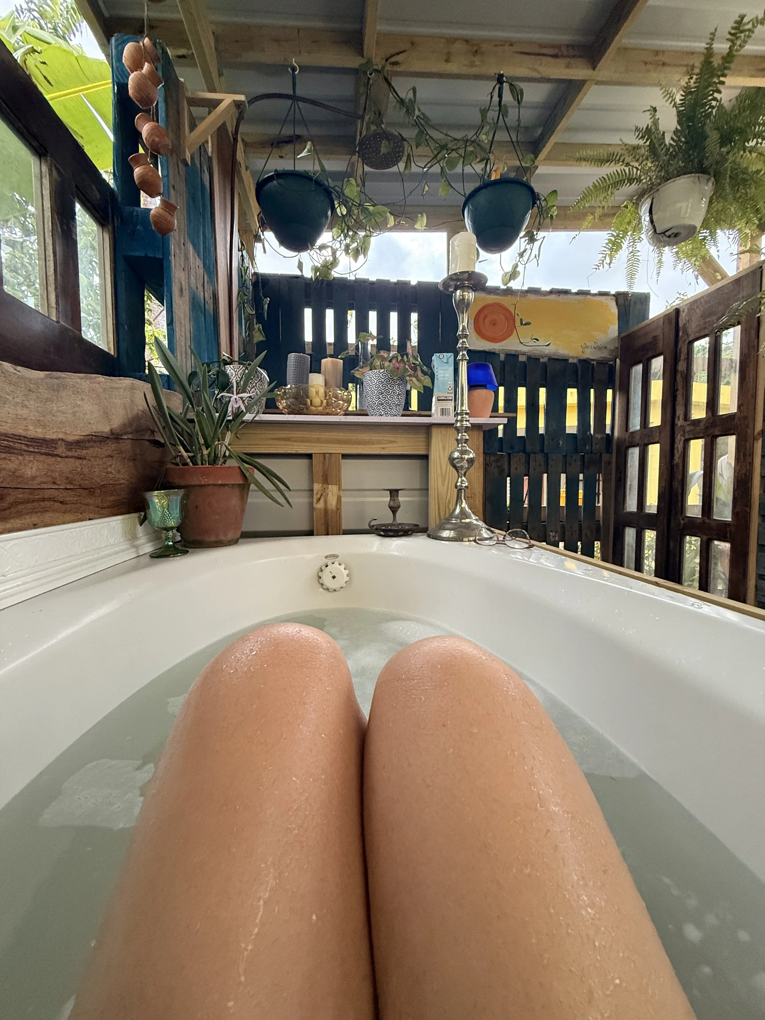 Person soaking in a bathtub with legs visible, surrounded by plants, candles, and decorative items in a cozy outdoor space.