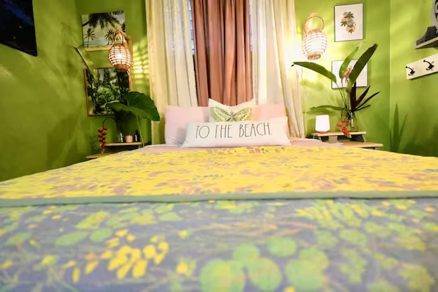 A bright bedroom with green walls, featuring a bed with floral yellow bedding, decorative pillows, and a "To the Beach" pillow. There are plants, wall art, and hanging lamps.