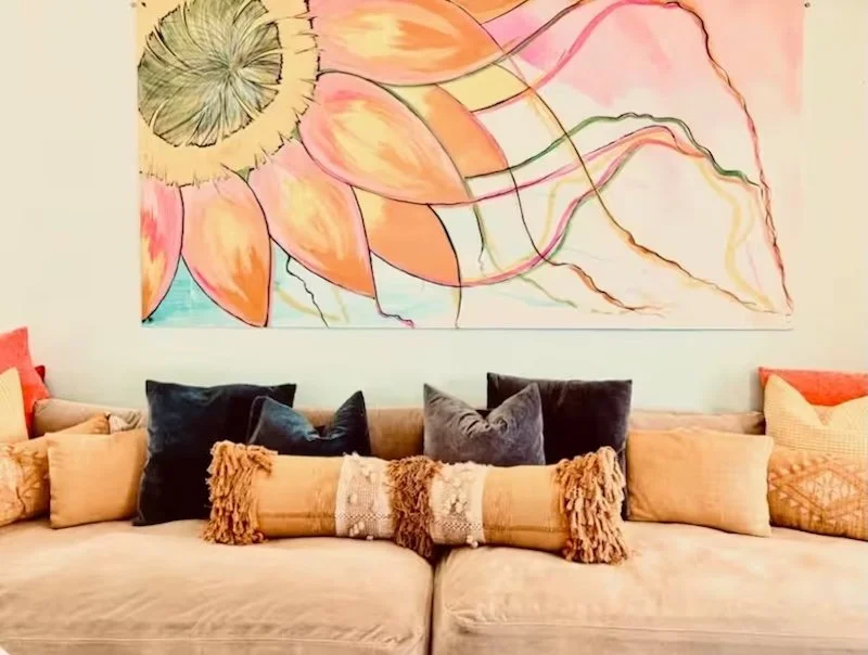 A cozy living room with a beige sofa adorned with various pillows in shades of black, beige, and orange. Hanging above the sofa is a large, colorful abstract floral painting with pink, orange, yellow, and green hues.