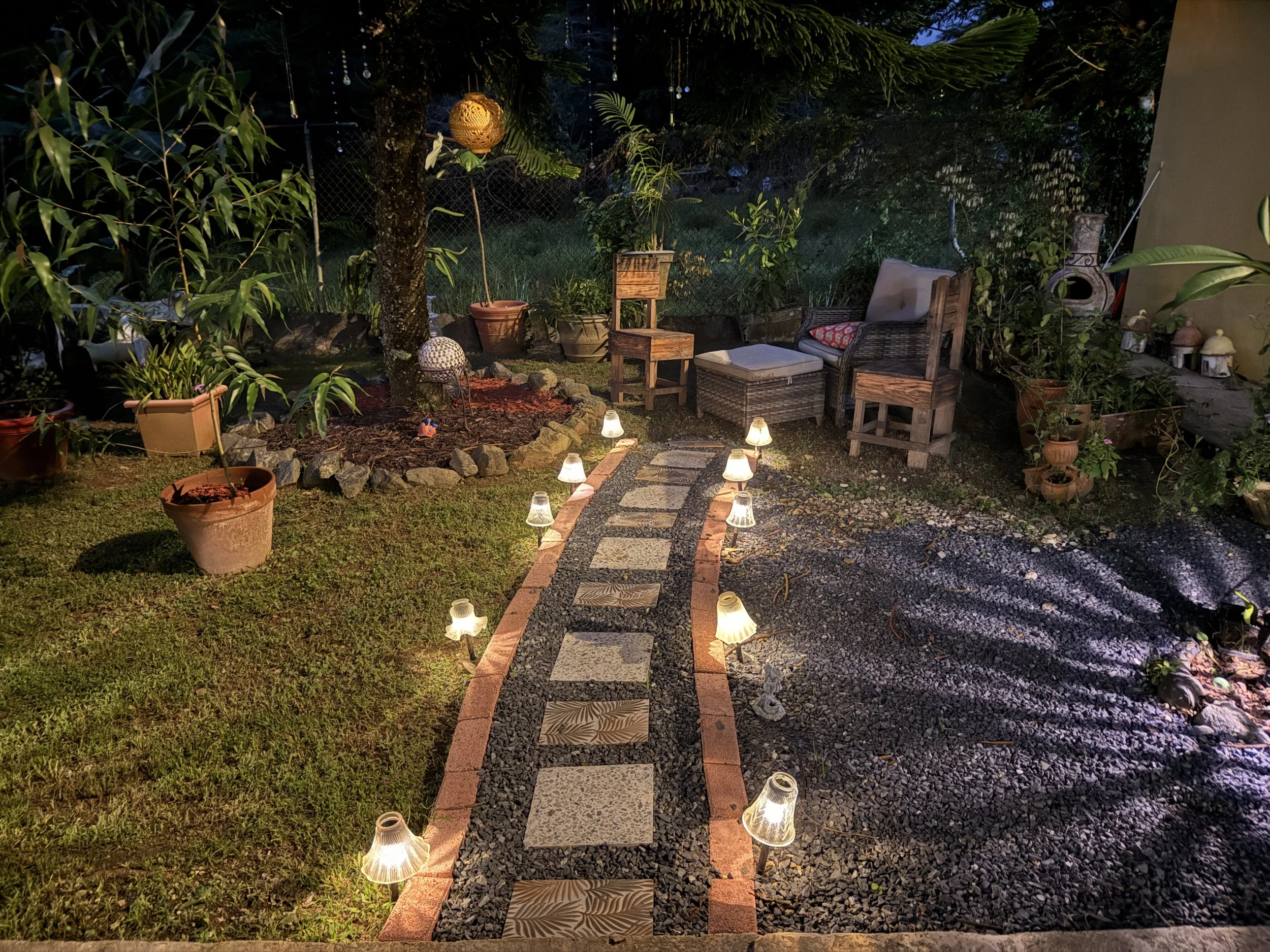 A backyard patio at night with a stone pathway lined by small solar-powered lights, surrounded by potted plants, outdoor furniture, and string lights hanging from trees.