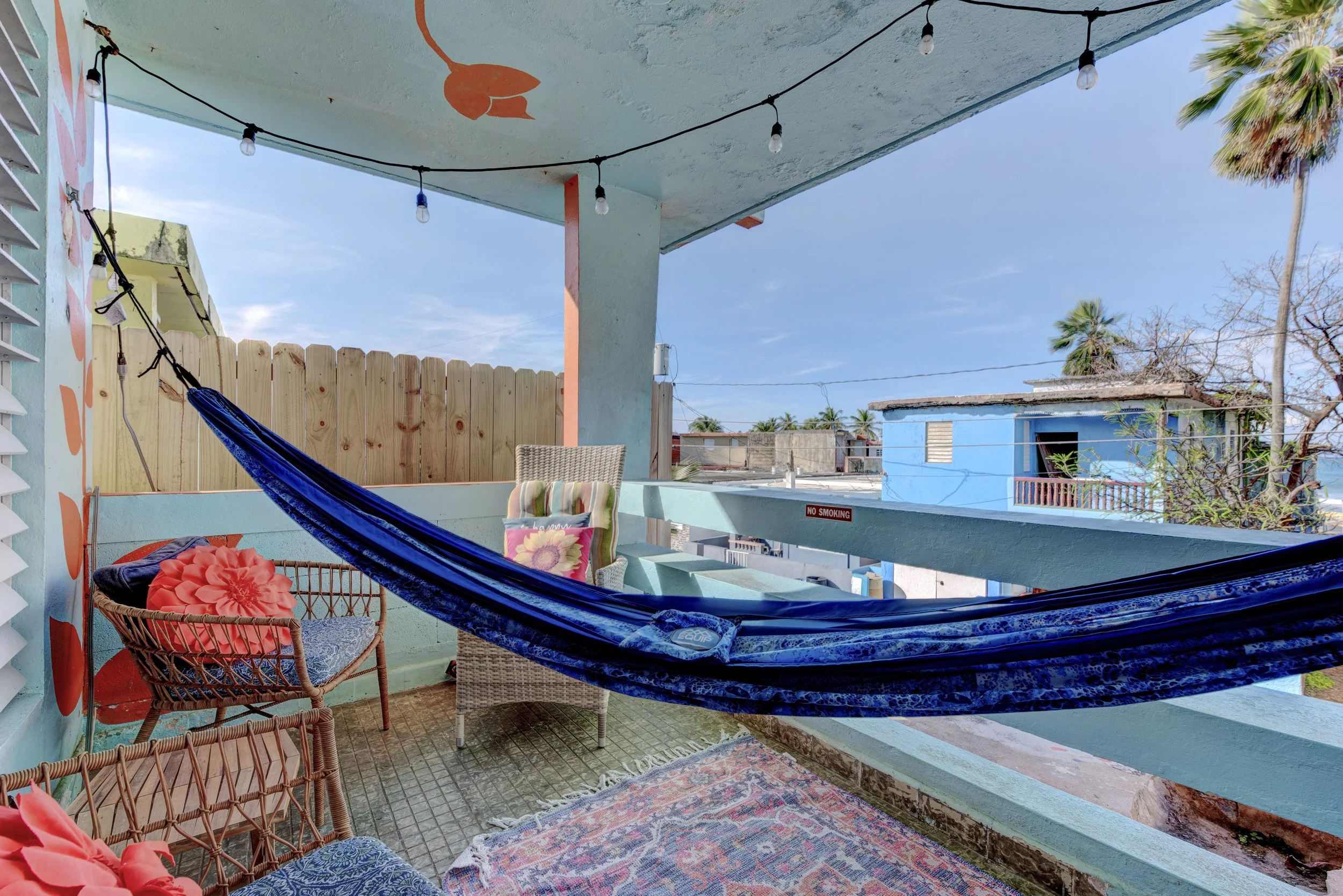 An apartment patio with colorful furniture, a hammock, string lights, and a clear sky with a few clouds, surrounded by a wooden fence and palm trees in the background.