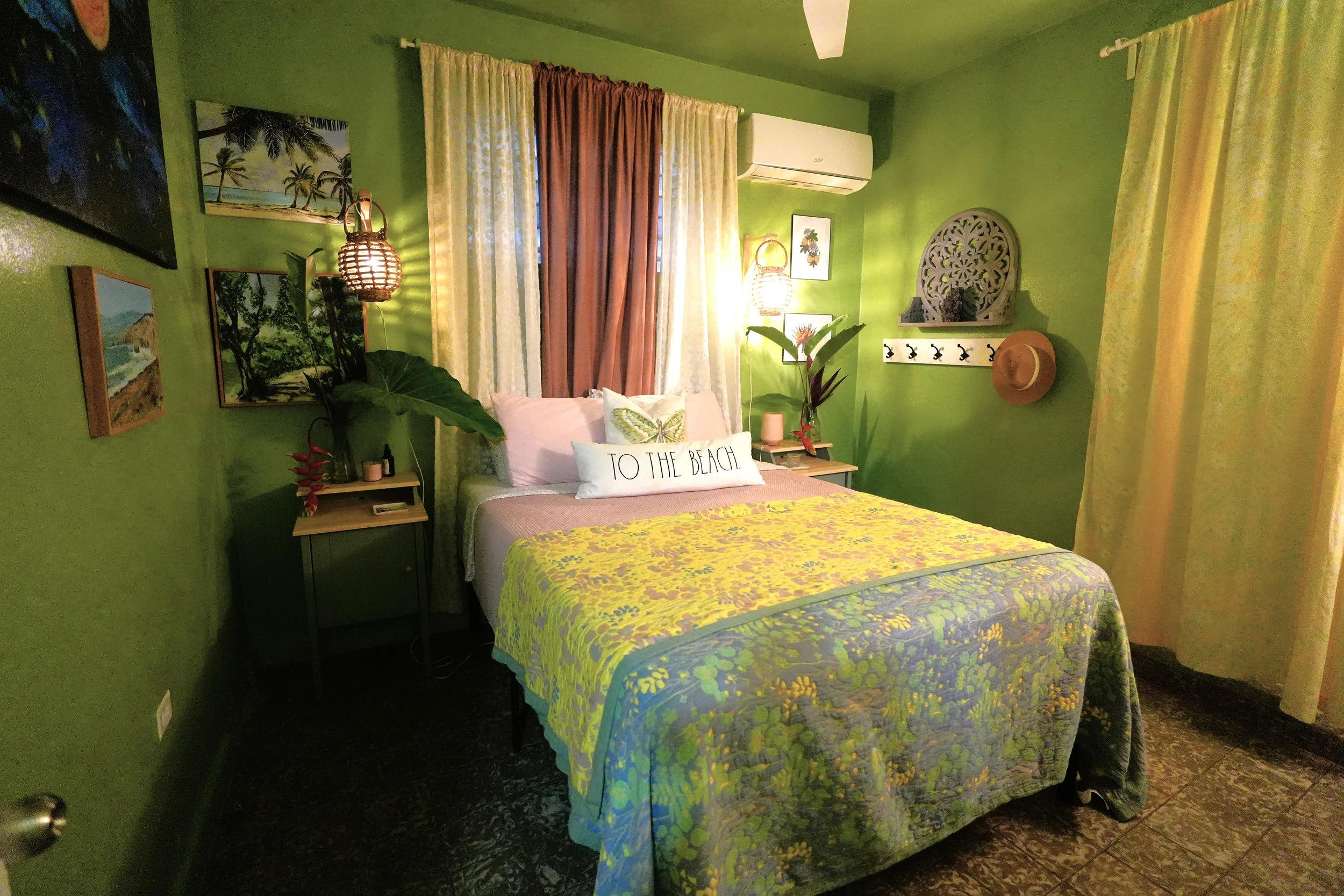 A bedroom with green walls and curtains, a bed with a yellow and green floral bedspread, tropical-themed artwork, and various decorative items including plants, lamps, and a straw hat.