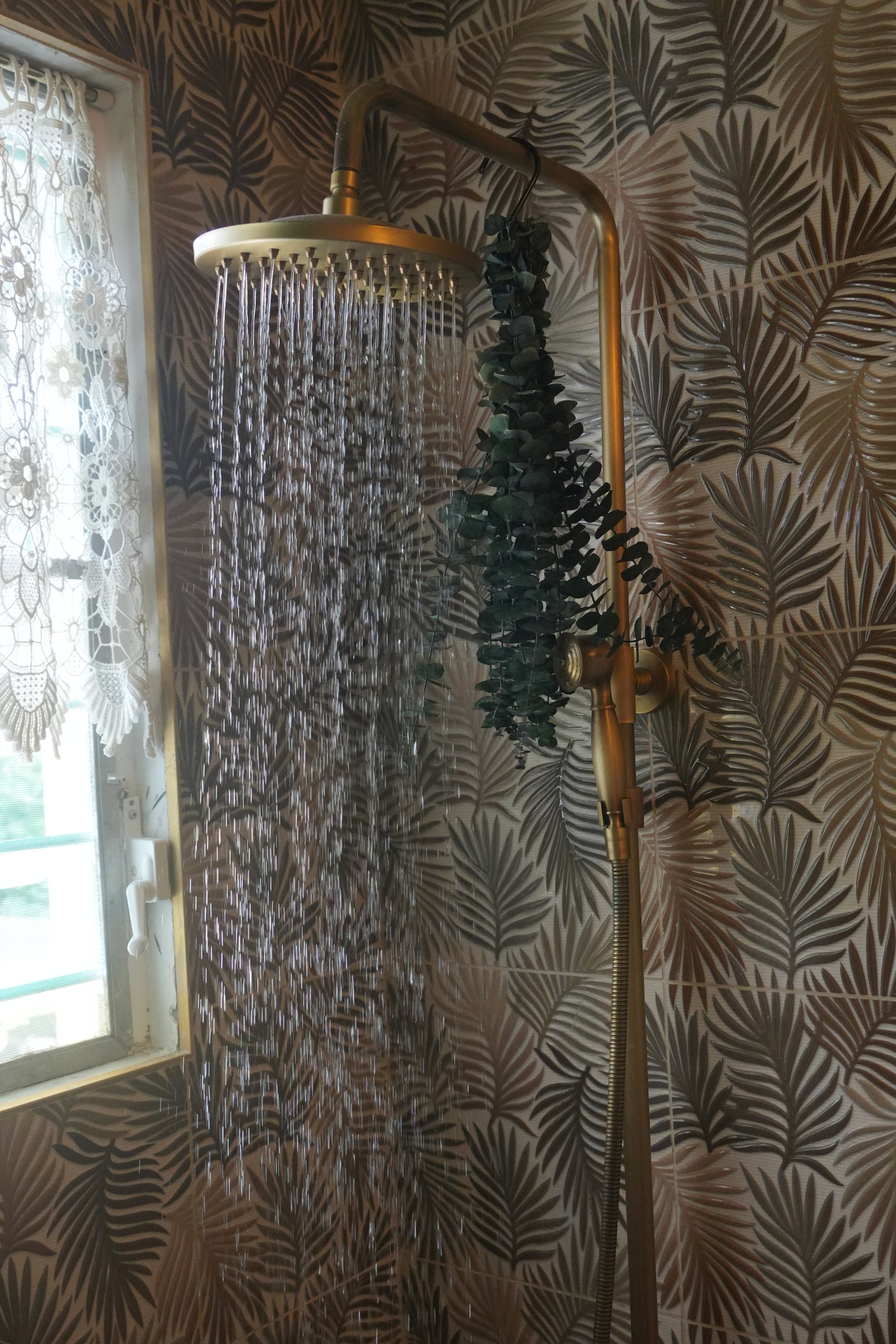 Shower with a gold-colored showerhead mounted on a decorative wall with leaf-patterned tile. Water is flowing from the showerhead. There is a window with lace curtains to the left.