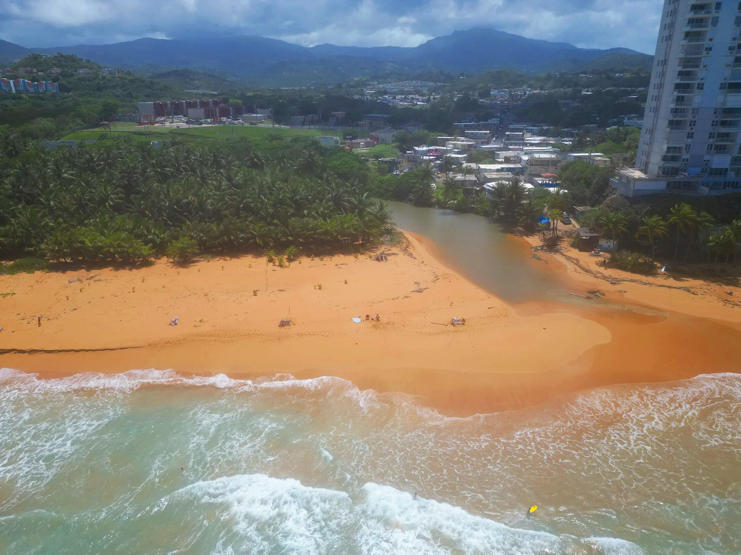 Costa Azul beach with golden sand, greenish ocean waves, and a small yellow boat near the shoreline. In the background, there is a lush green area with palm trees, residential buildings, and mountains under a cloudy sky.