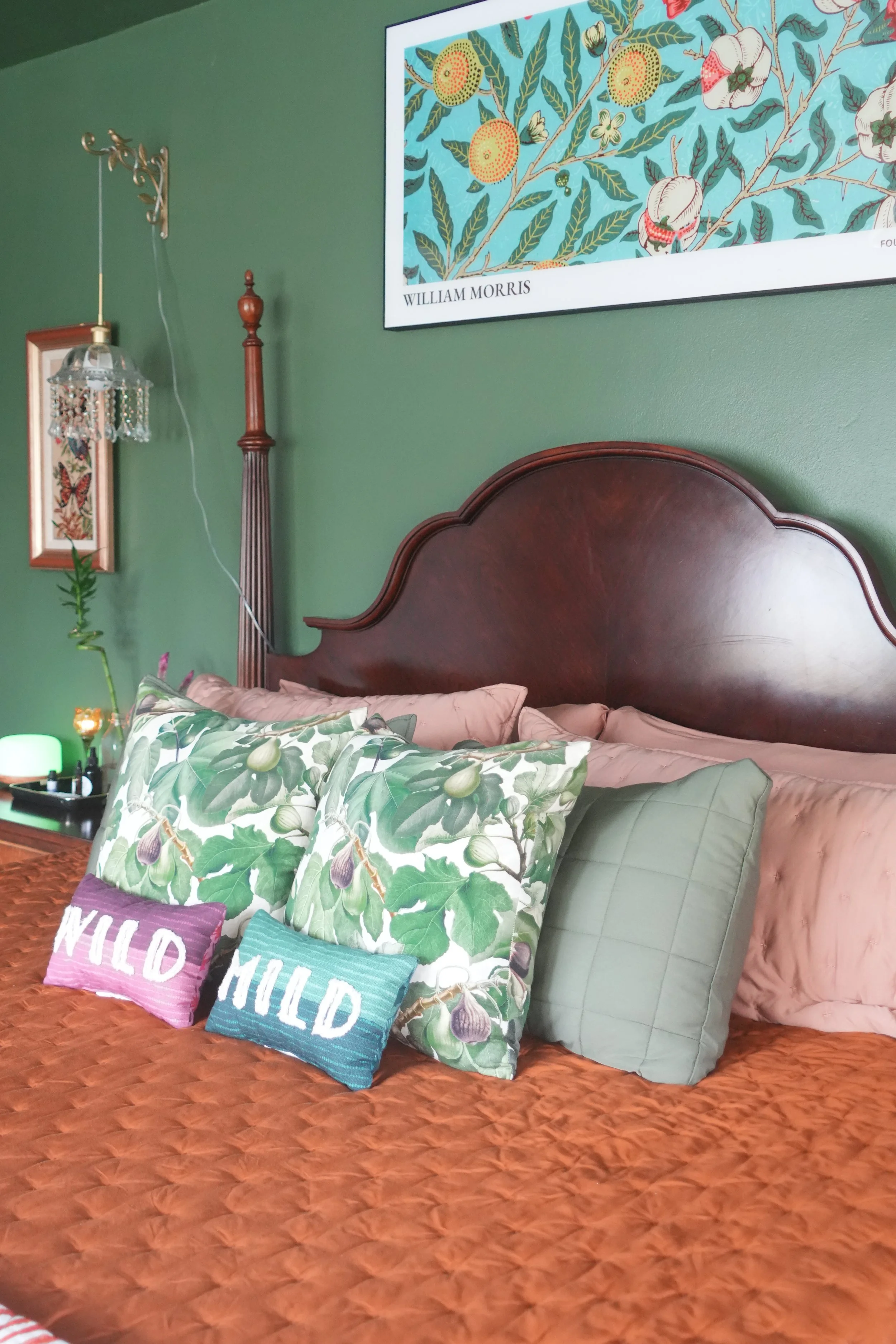 A bedroom with a green wall and wooden bed frame. On the bed, there are pillows with botanical patterns, including fig leaves and figs. Above the bed, there is a large framed artwork by William Morris featuring botanical illustrations. To the left, t