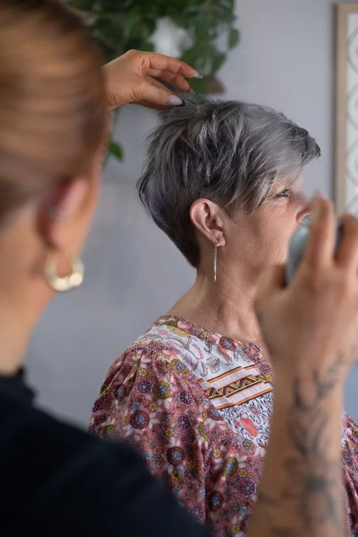A hairstylist styling a senior woman's short gray hair in a salon. The woman is wearing a floral top and earrings, and the stylist's hand is visible in the foreground.