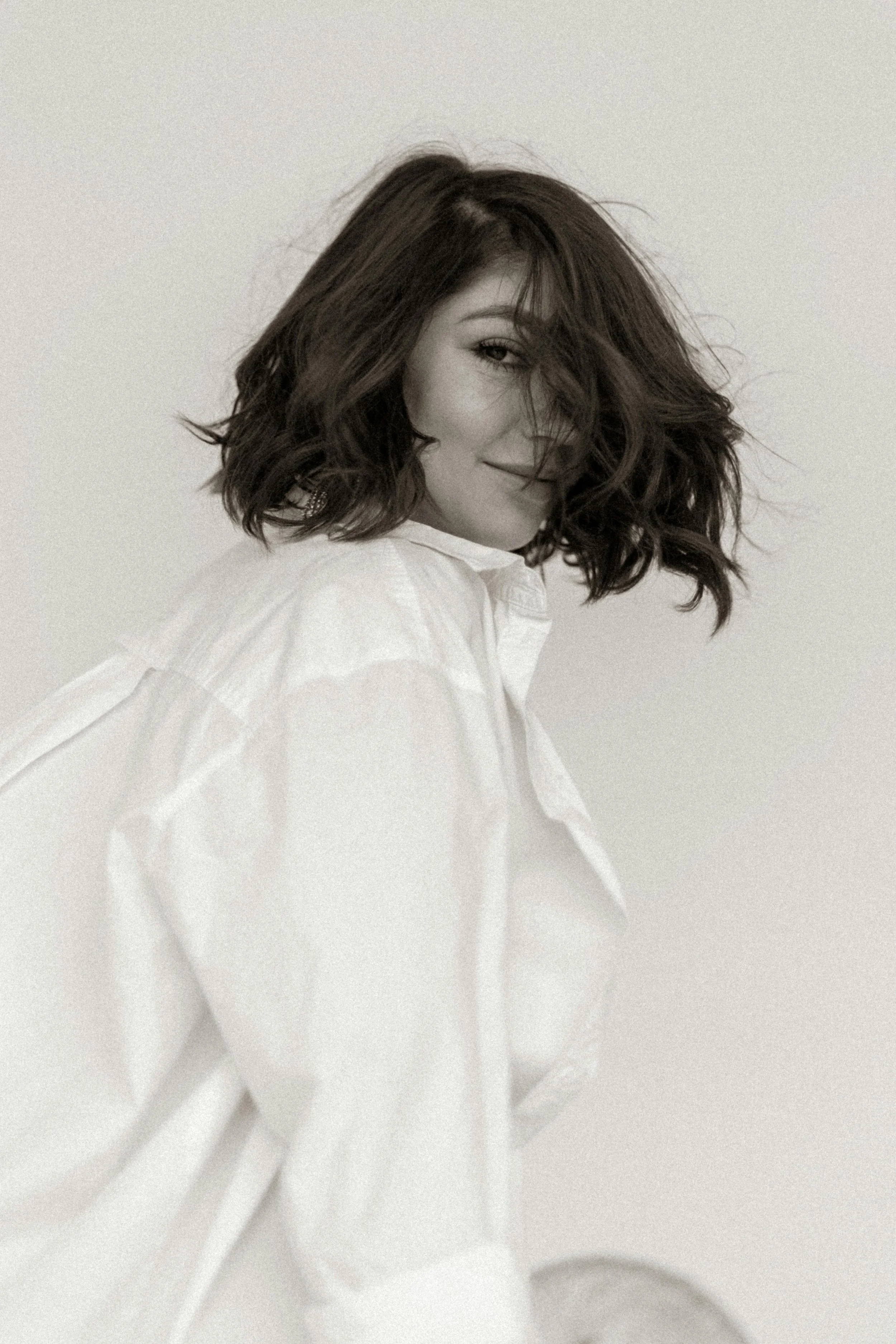 A woman with shoulder-length wavy hair, wearing a white shirt, smiling with her face partially turned and hair covering one eye.