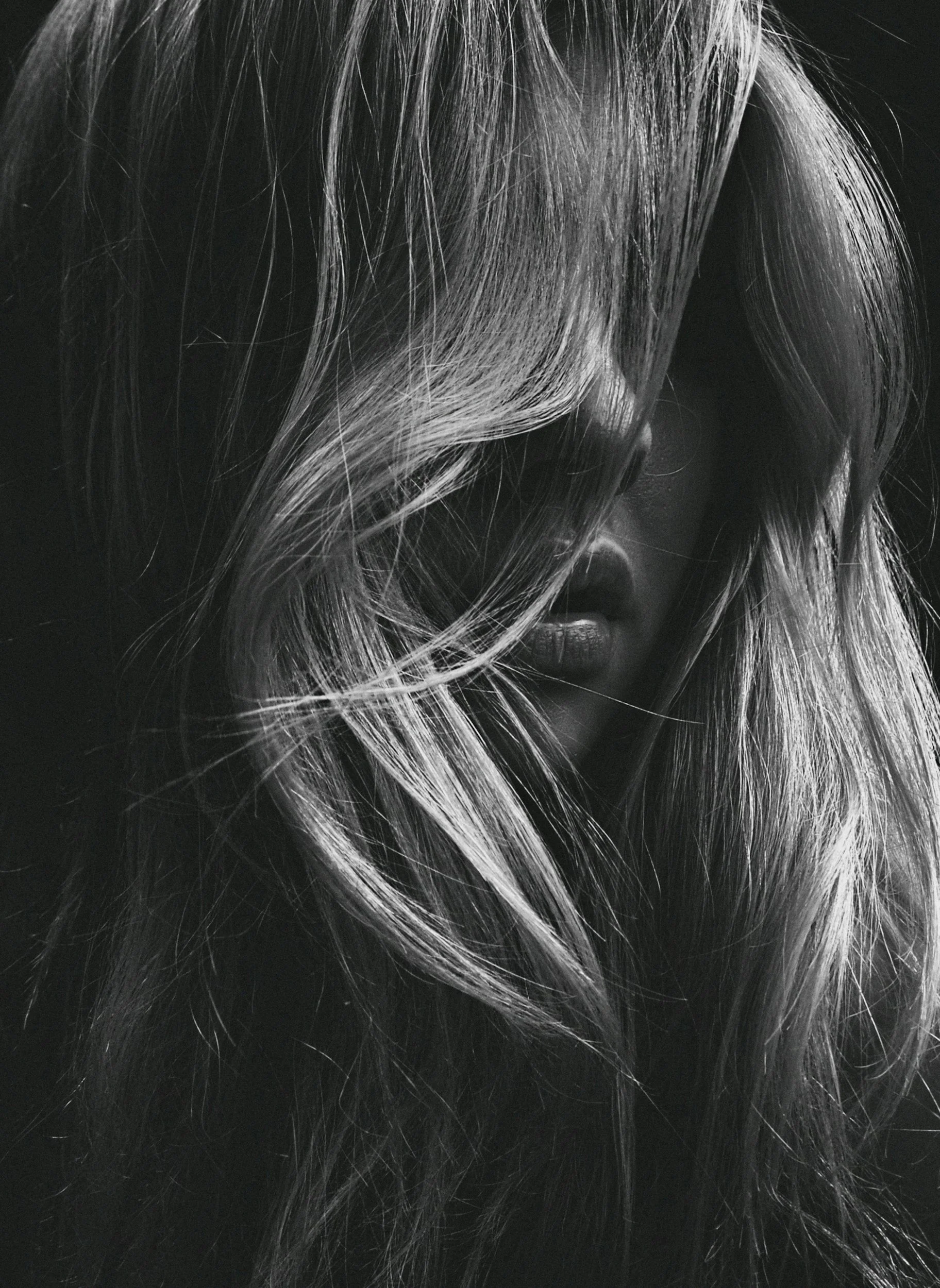 A black-and-white photograph of a woman with voluminous, wavy hair partially covering her face, with visible lips and nose.