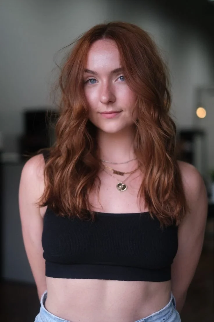 A young woman with long, wavy red hair and blue eyes, wearing a black crop top and layered necklaces, standing in a softly lit indoor setting.