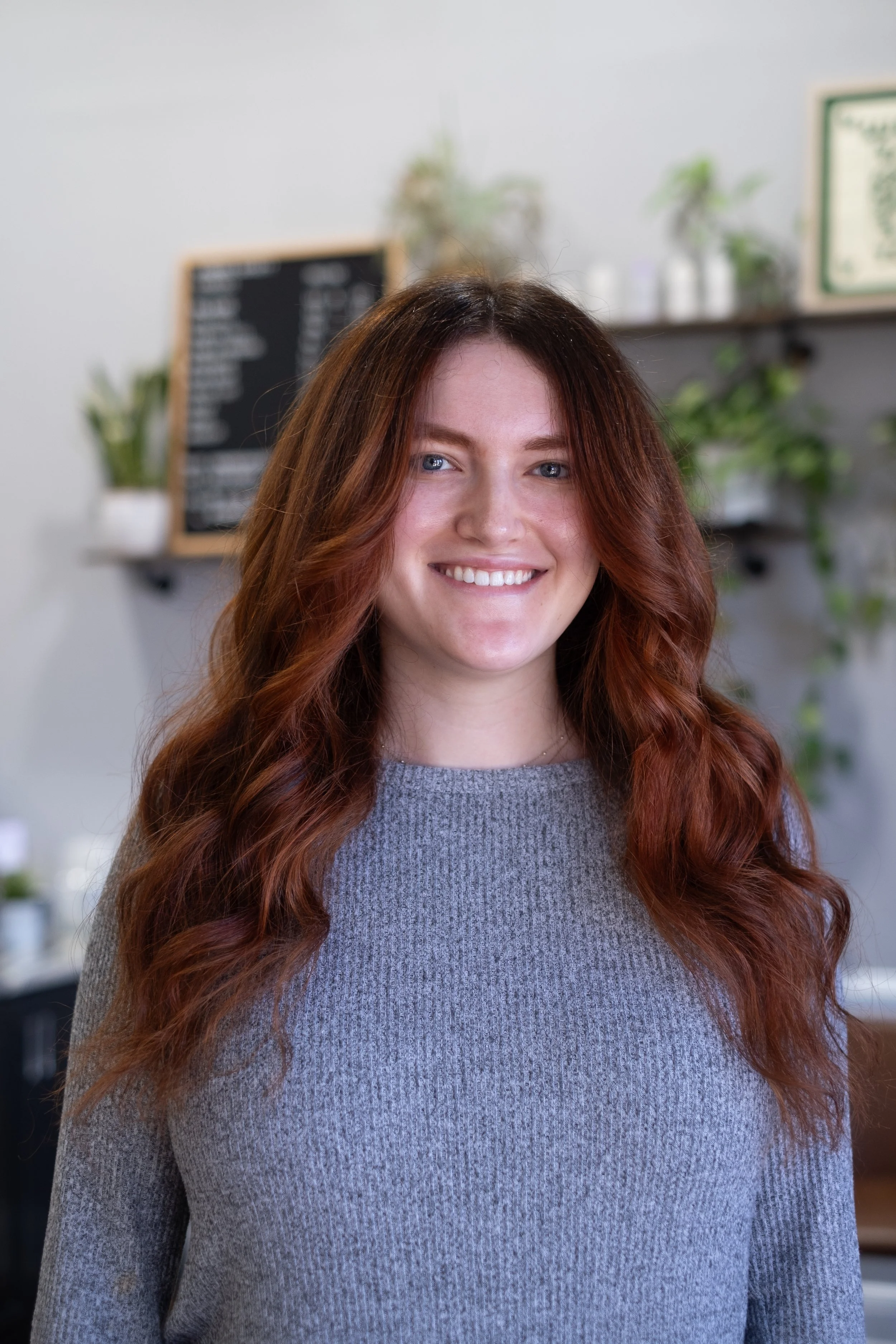 A young woman with long, wavy red hair and a gray sweater, smiling at the camera inside a cafe with a blackboard menu and potted plants in the background.